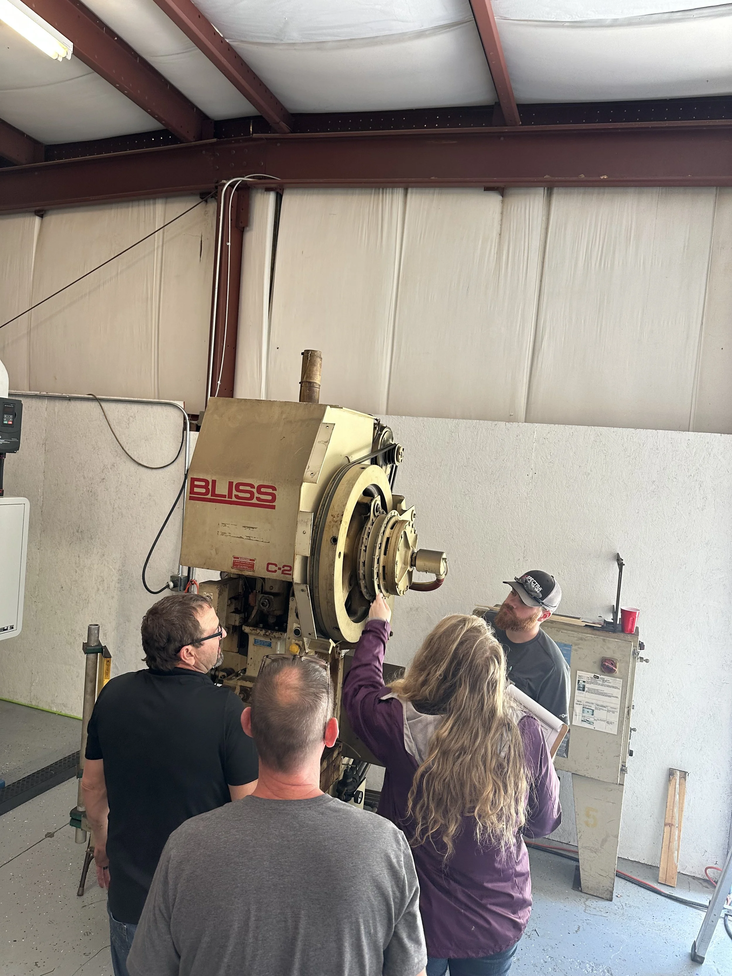 Four people are gathered around a large industrial machine labeled 'BLISS', with a woman pointing at it, inside a workshop or factory setting.