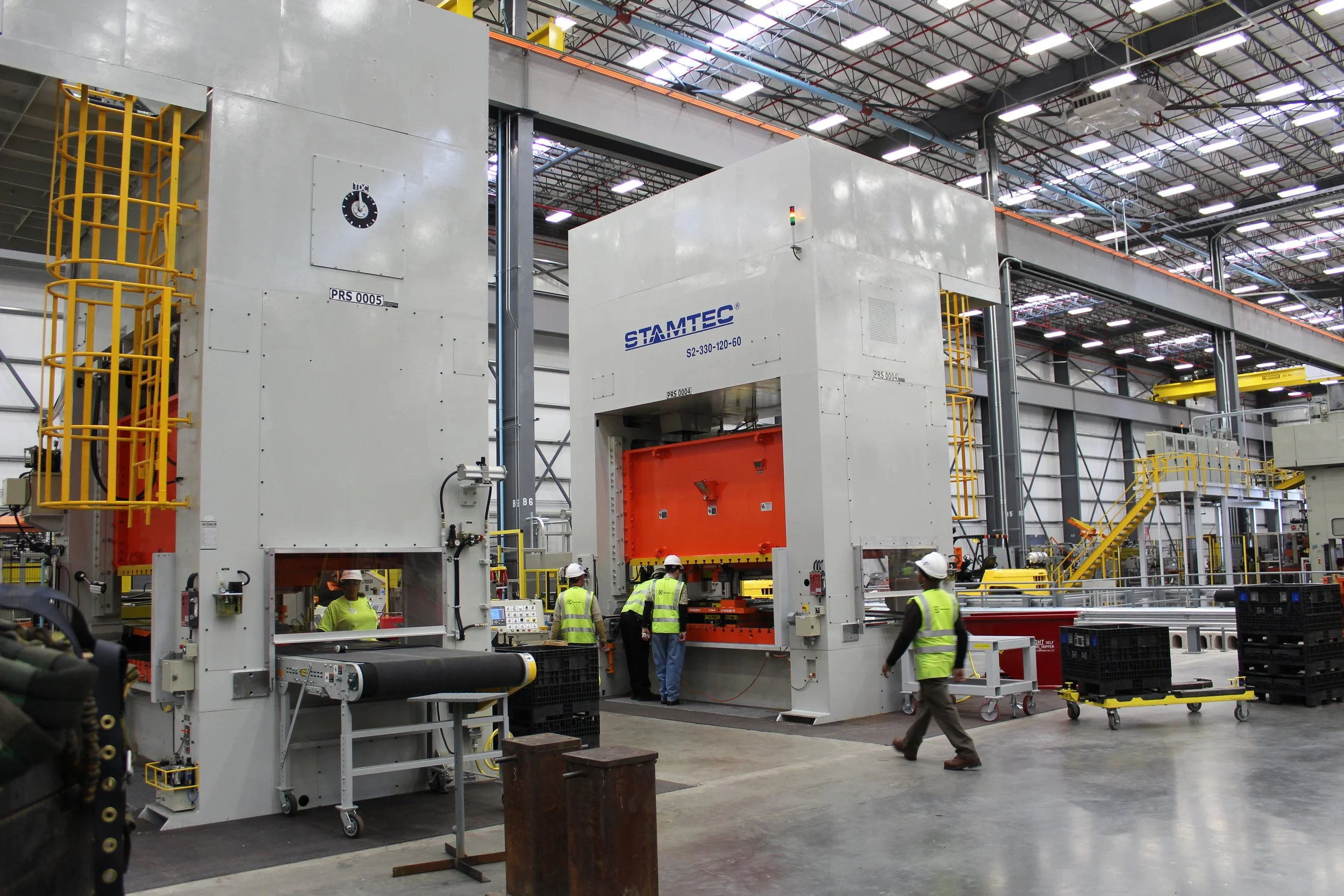 Workers in safety vests and helmets operate large industrial machinery inside a spacious factory with high ceilings and yellow safety barriers.