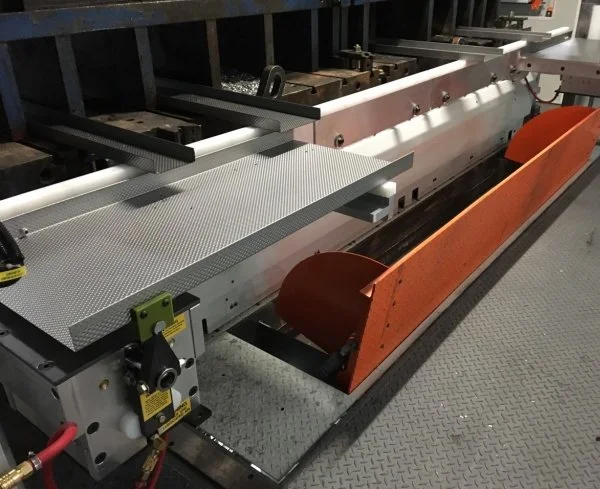 Industrial metal fabrication machine with metal work surface, orange safety guard, and control panel in a workshop.