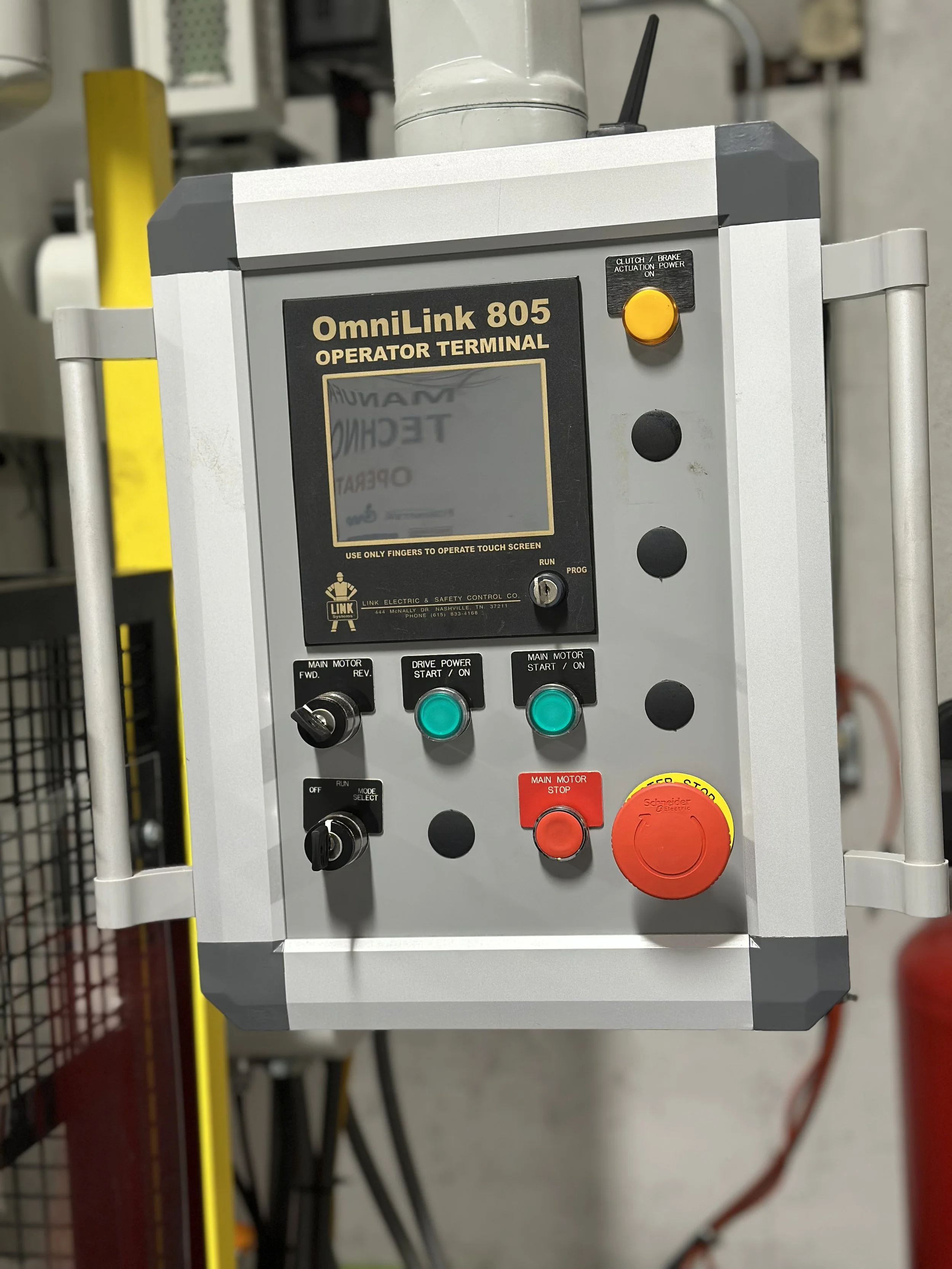 A control panel labeled 'OmniLink 805 Operator Terminal' with buttons and switches, including green start buttons, a red emergency stop button, and black knobs, mounted on a wall in an industrial setting.