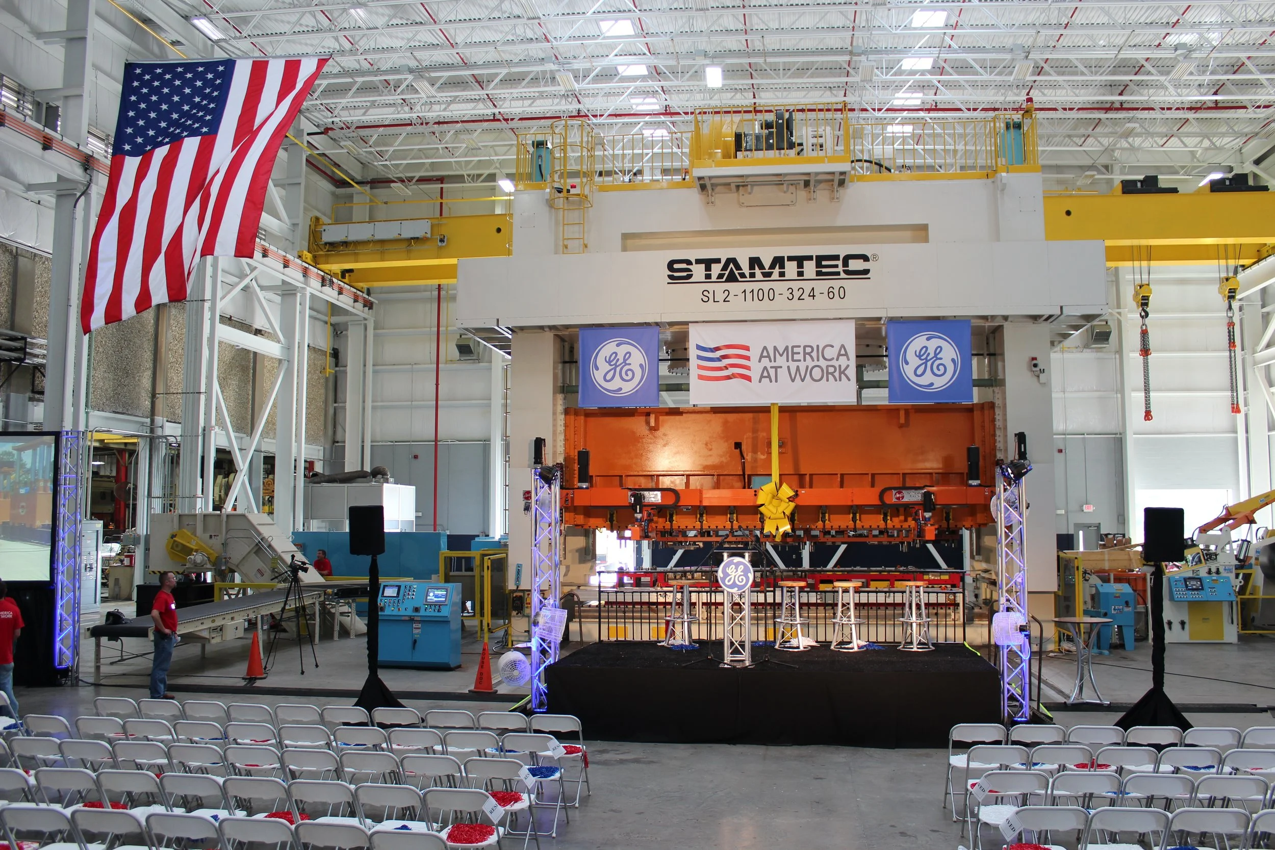 Inside a large industrial facility with a stage set up for an event, featuring a backdrop with the GE logo, American flag, and the slogan 'America at Work.' There are rows of empty chairs in the foreground and large industrial equipment and machinery