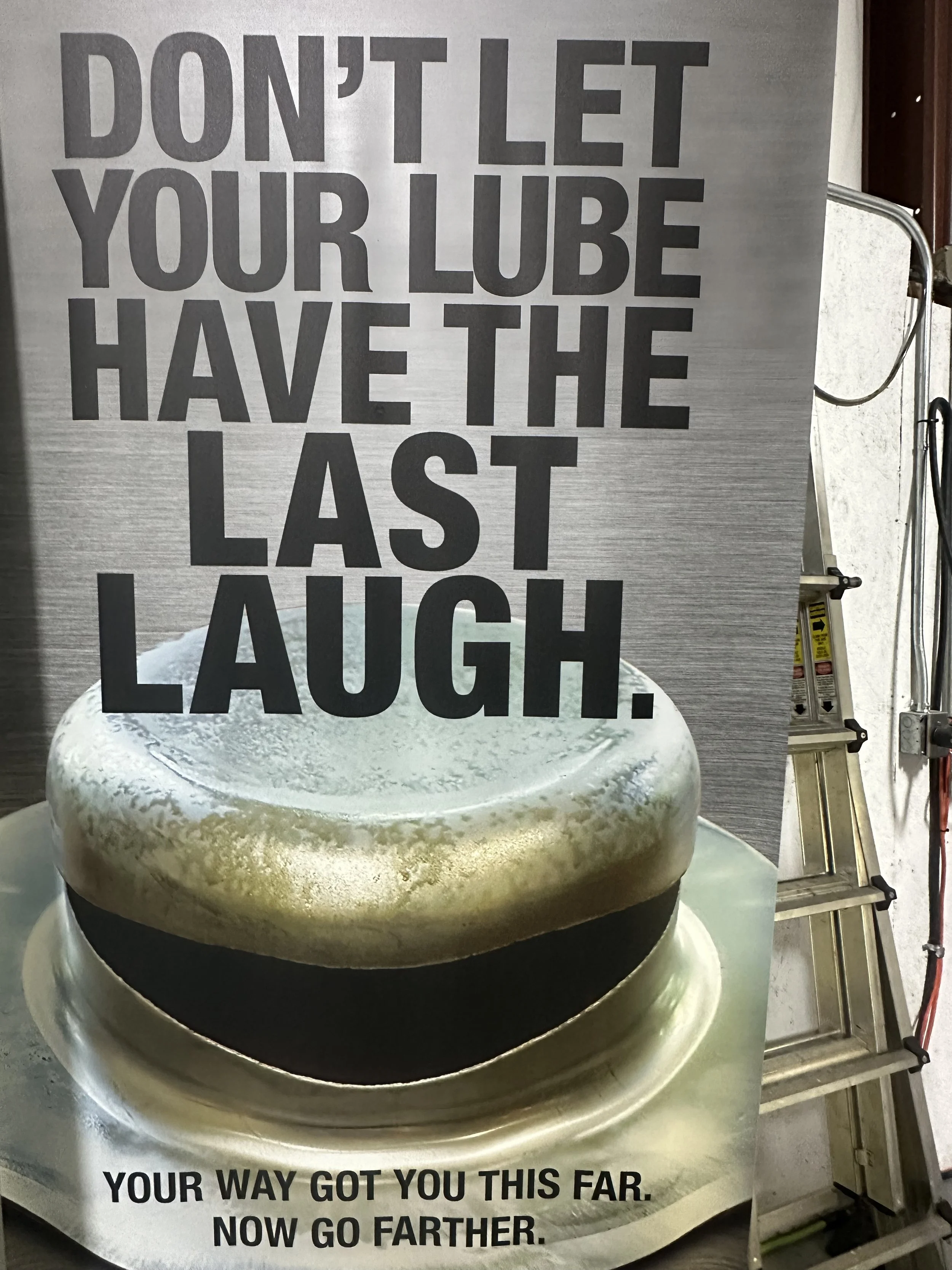 A sign with a humorous quote about lube, featuring an image of a large lubricant container, with the text: "DON'T LET YOUR LUBE HAVE THE LAST LAUGH. YOUR WAY GOT YOU THIS FAR. NOW GO FARTHER."
