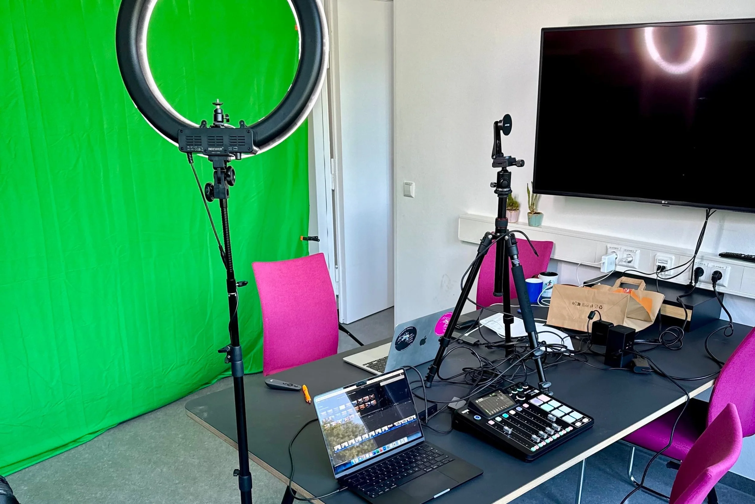 Indoor setup for a video or photo shoot with a green screen, two pink chairs, laptops, a camera on a tripod, lighting equipment, and various electronic accessories on a table.