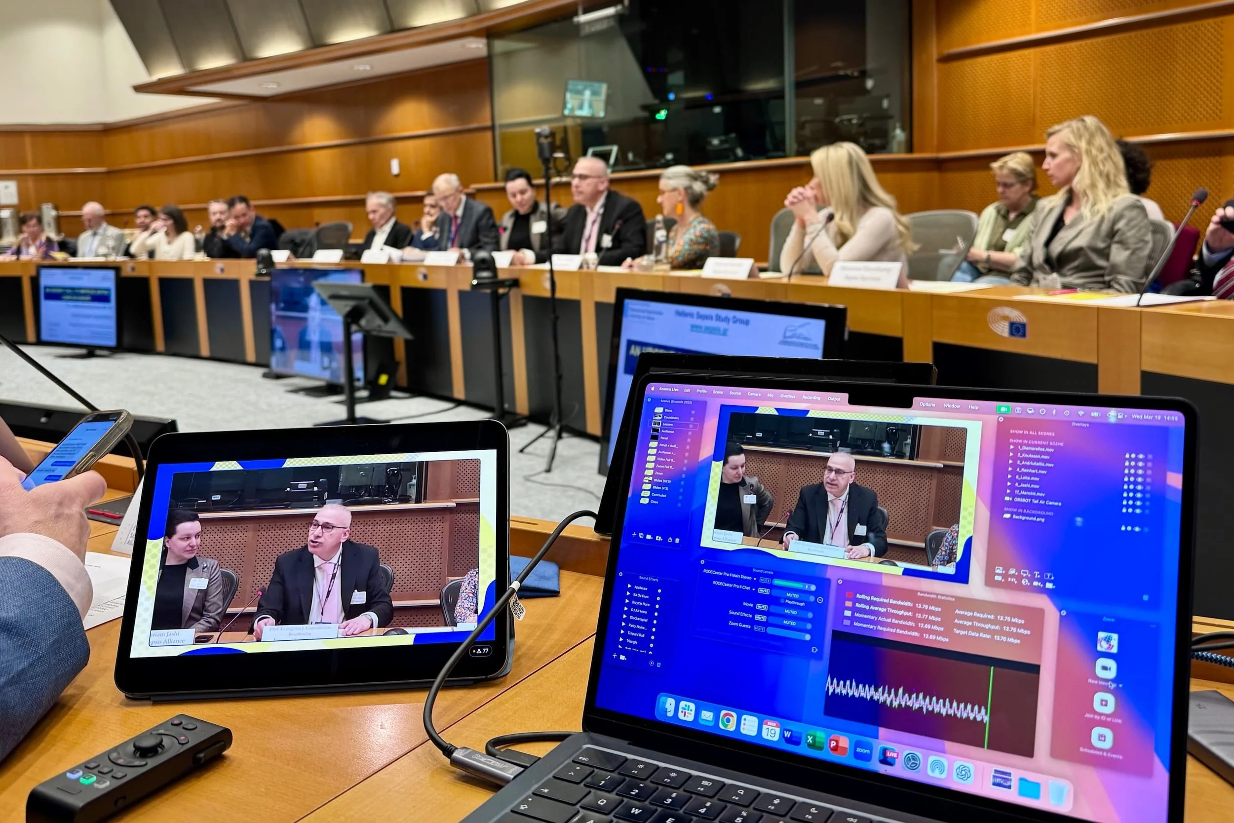 Conference room with a long table, attendees seated on both sides, and multiple electronic screens displaying a panel discussion in progress.