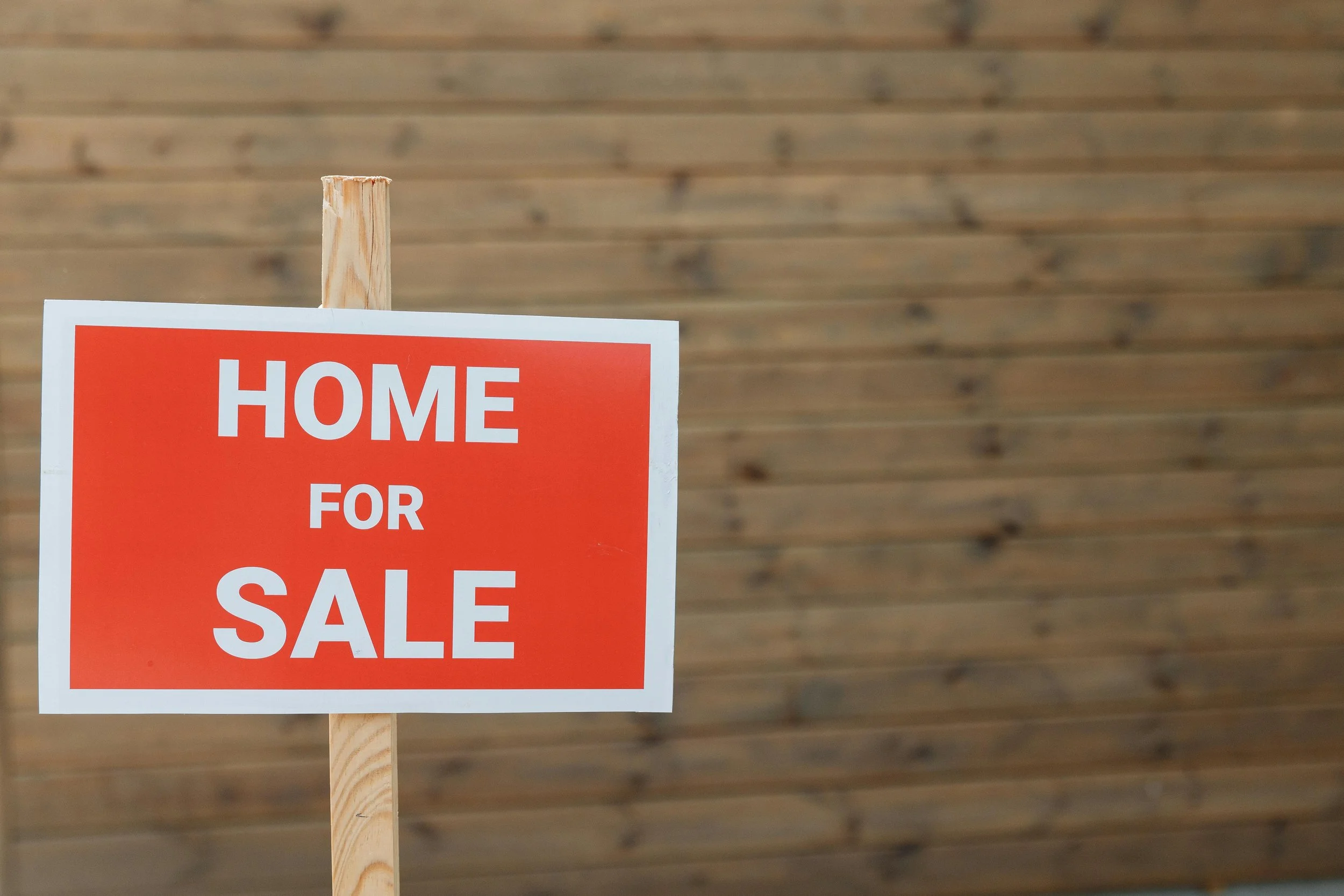 A sign with a red background and white text that reads 'Home for Sale' is attached to a wooden post. The background consists of a wooden wall.