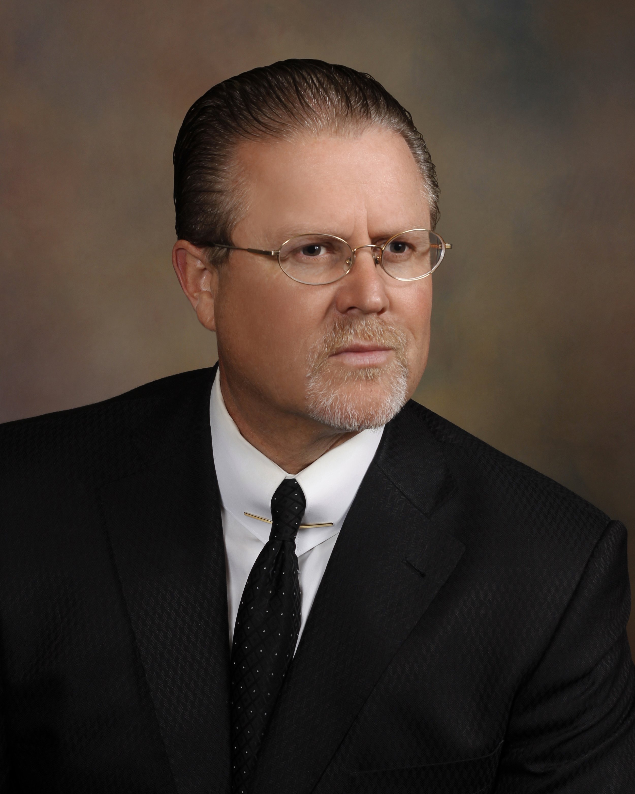 A middle-aged man with slicked-back hair, glasses, and a beard, wearing a black suit, white shirt, and black tie, looking directly at the camera against a neutral background.