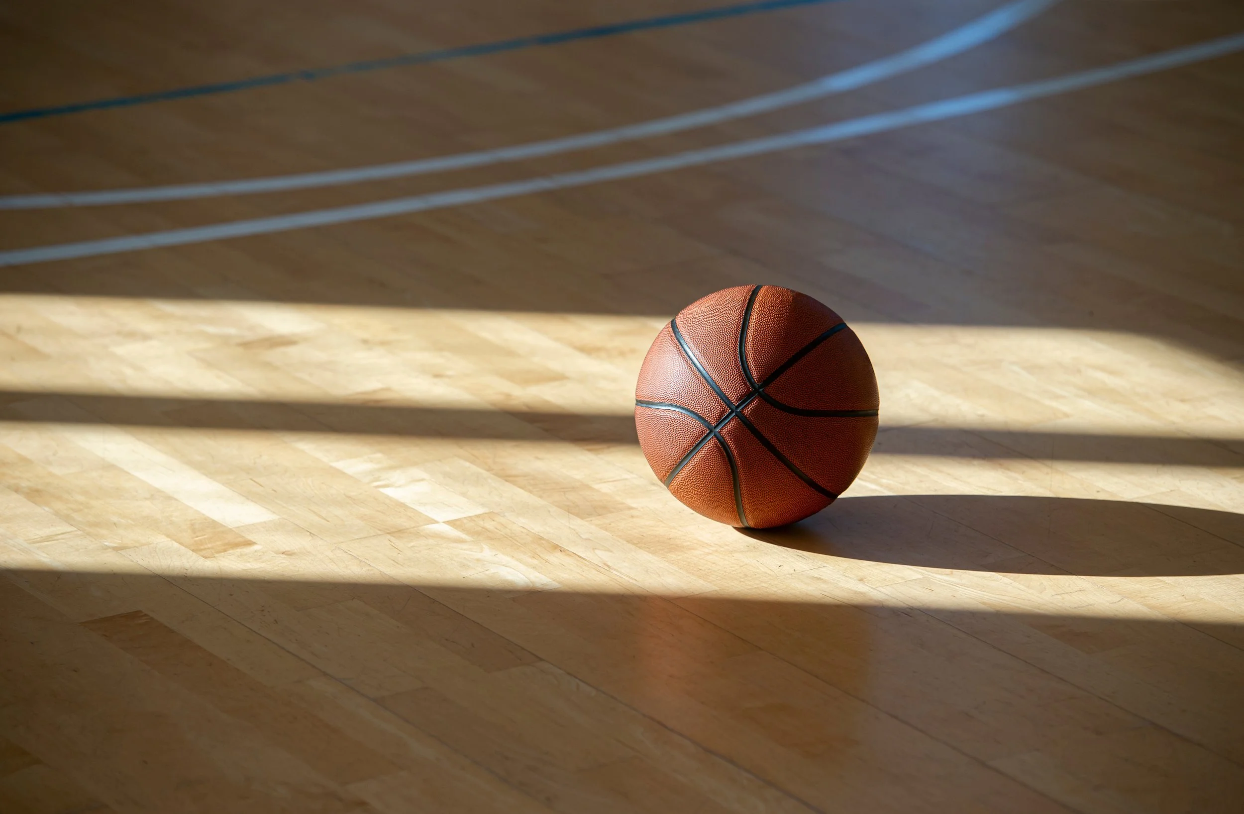 A basketball on a wooden gym court floor with sunlight creating shadows.