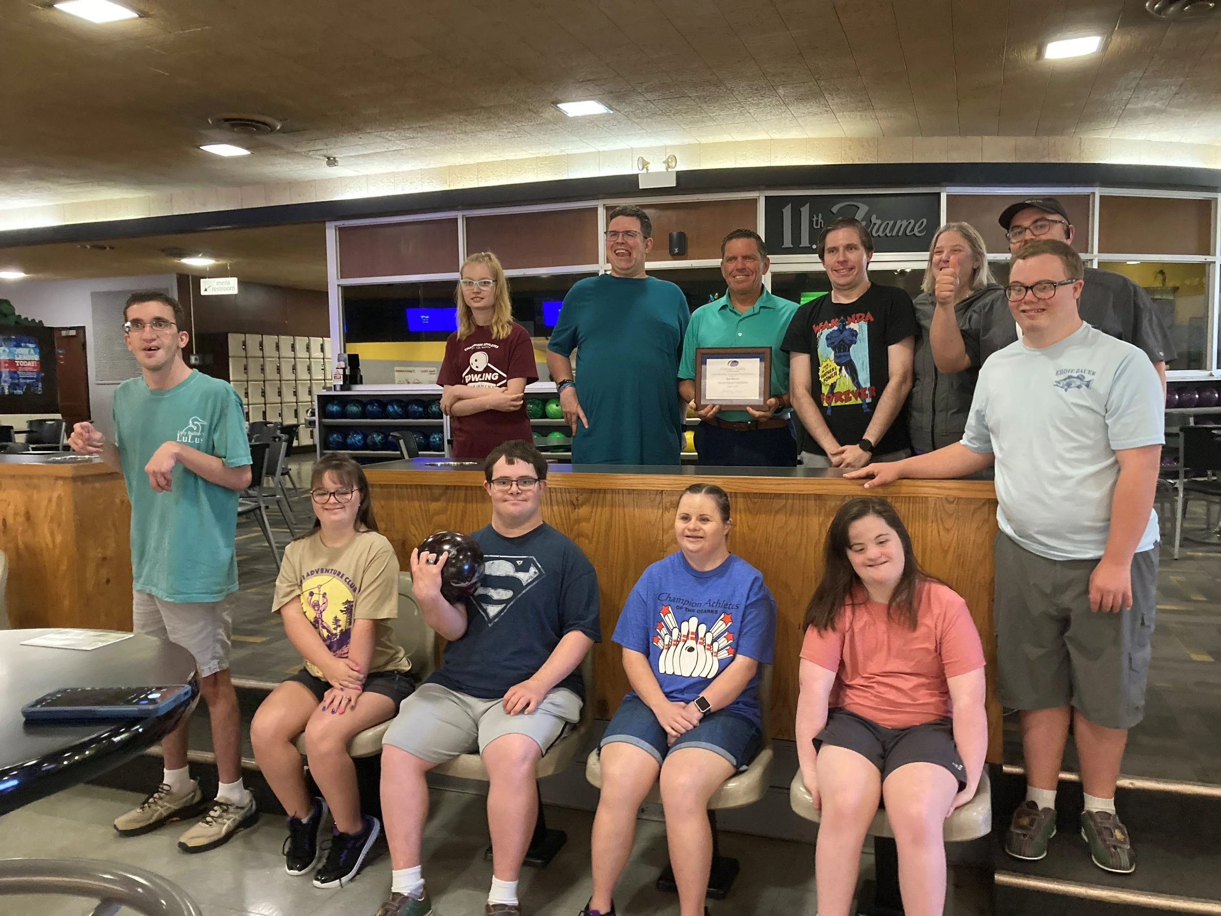 A group of children and adults at a bowling alley, posing for a photo. Some children are seated, others are standing, and one adult is holding a framed certificate. The background shows bowling balls and a sign that reads '11th Frame.'