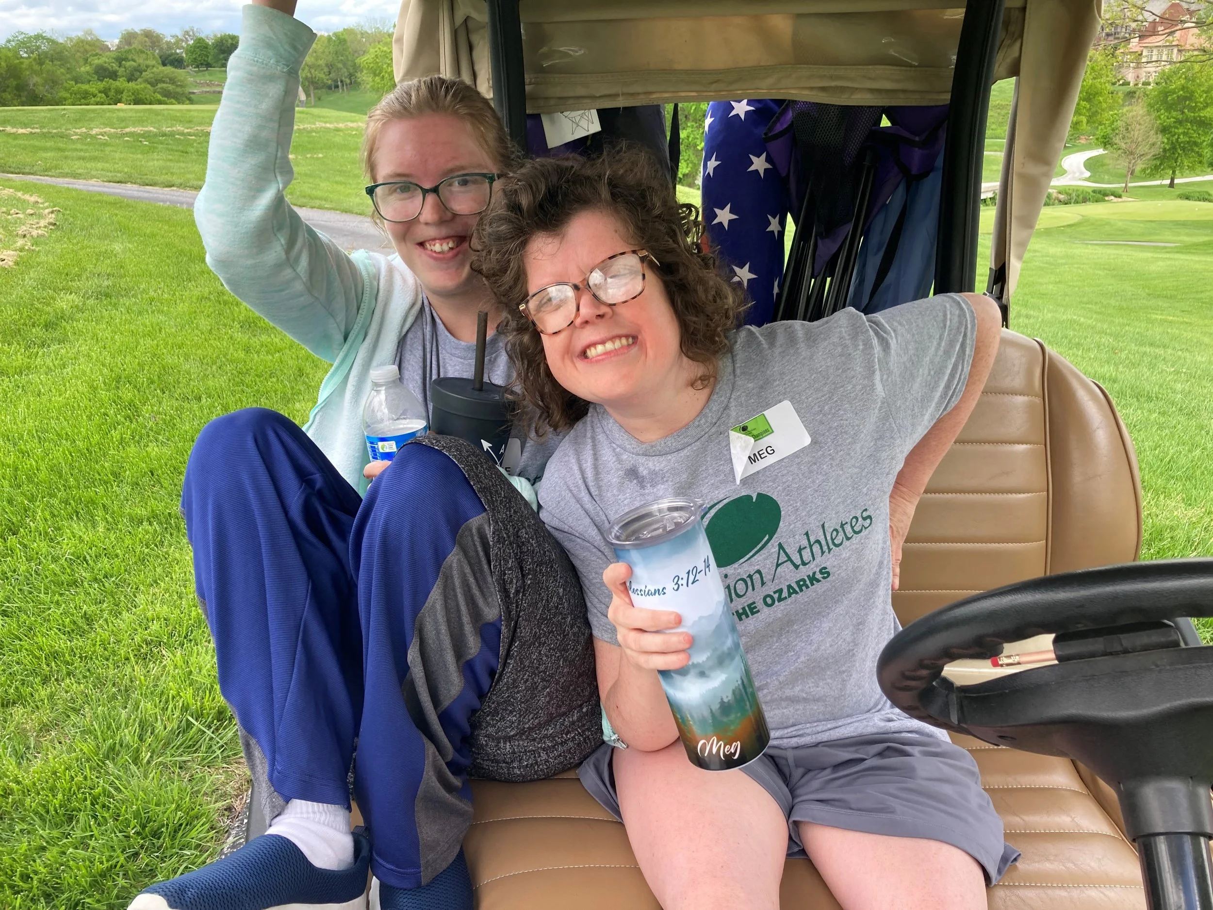 Two women sitting on a golf cart, smiling and enjoying time outdoors on a golf course with green grass and trees in the background. One woman has curly hair, glasses, and a gray shirt, while the other woman has straight hair, glasses, and a light blue jacket. They are holding drinks, and there is golf equipment behind them.