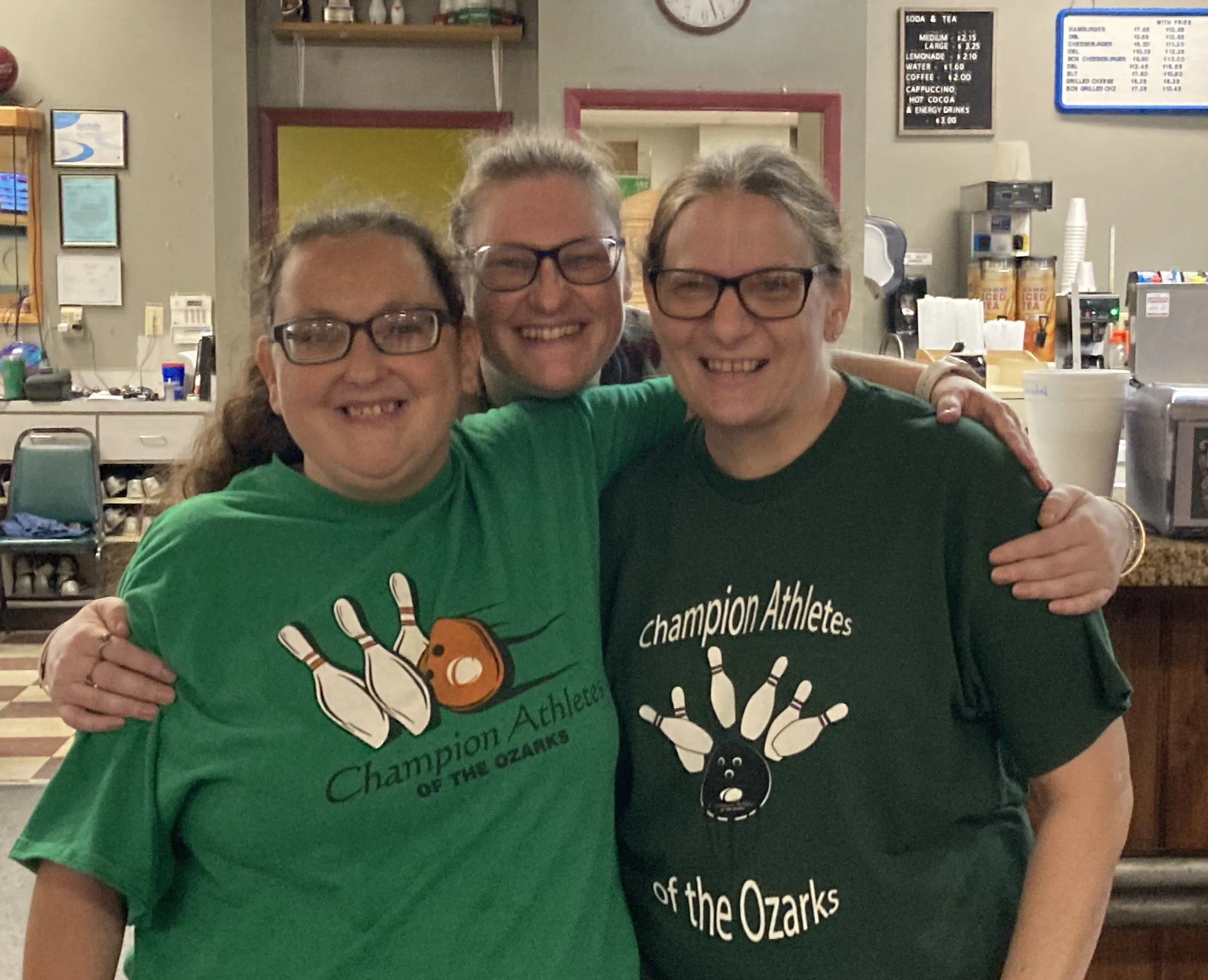 Three smiling women with glasses, wearing green and black shirts, hugging each other inside a cafe or restaurant.