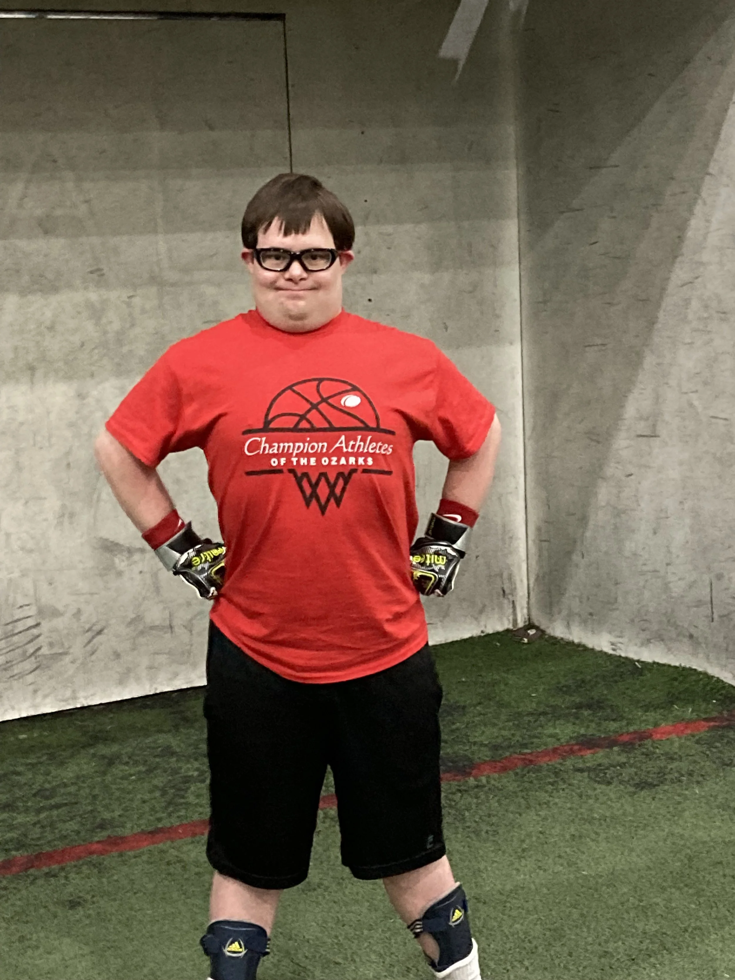 A young man in a red sports shirt with 'Champion Athletes of the Ozarks' printed on it, wearing black shorts, knee pads, and gloves, standing on an indoor sports turf field with a concrete wall in the background.