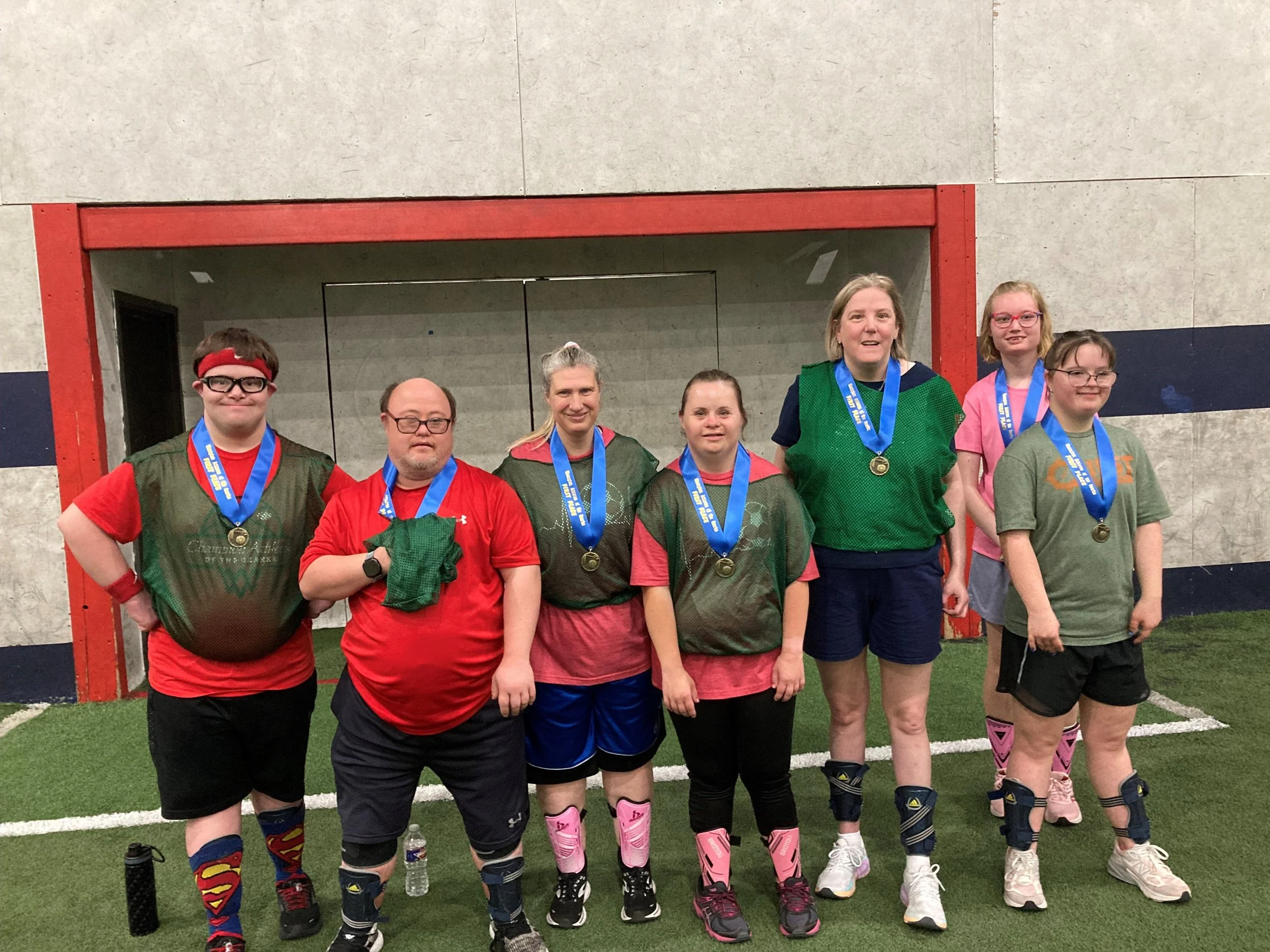 Group of six women and one man wearing sports gear and medals, standing inside an indoor sports facility.