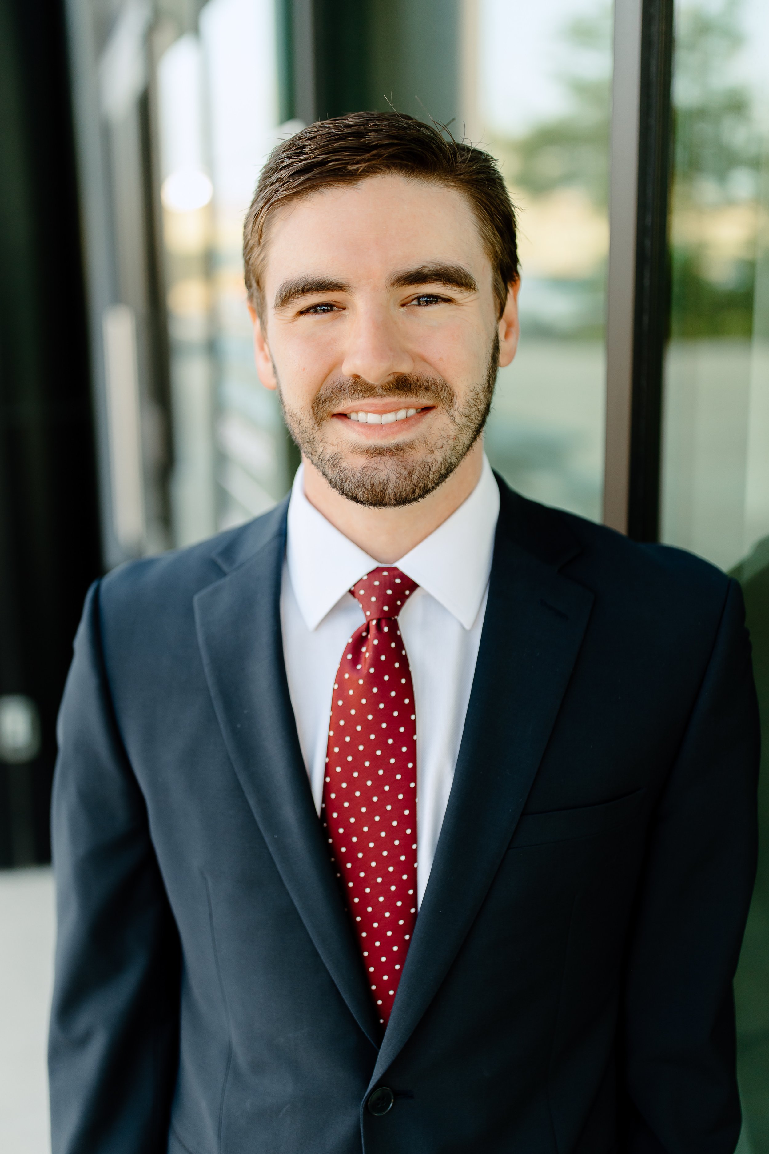 A young man with brown hair, beard, and blue eyes, dressed in a navy suit, white shirt, and red polka dot tie, standing indoors near a large window with blurred outdoor scenery in the background.