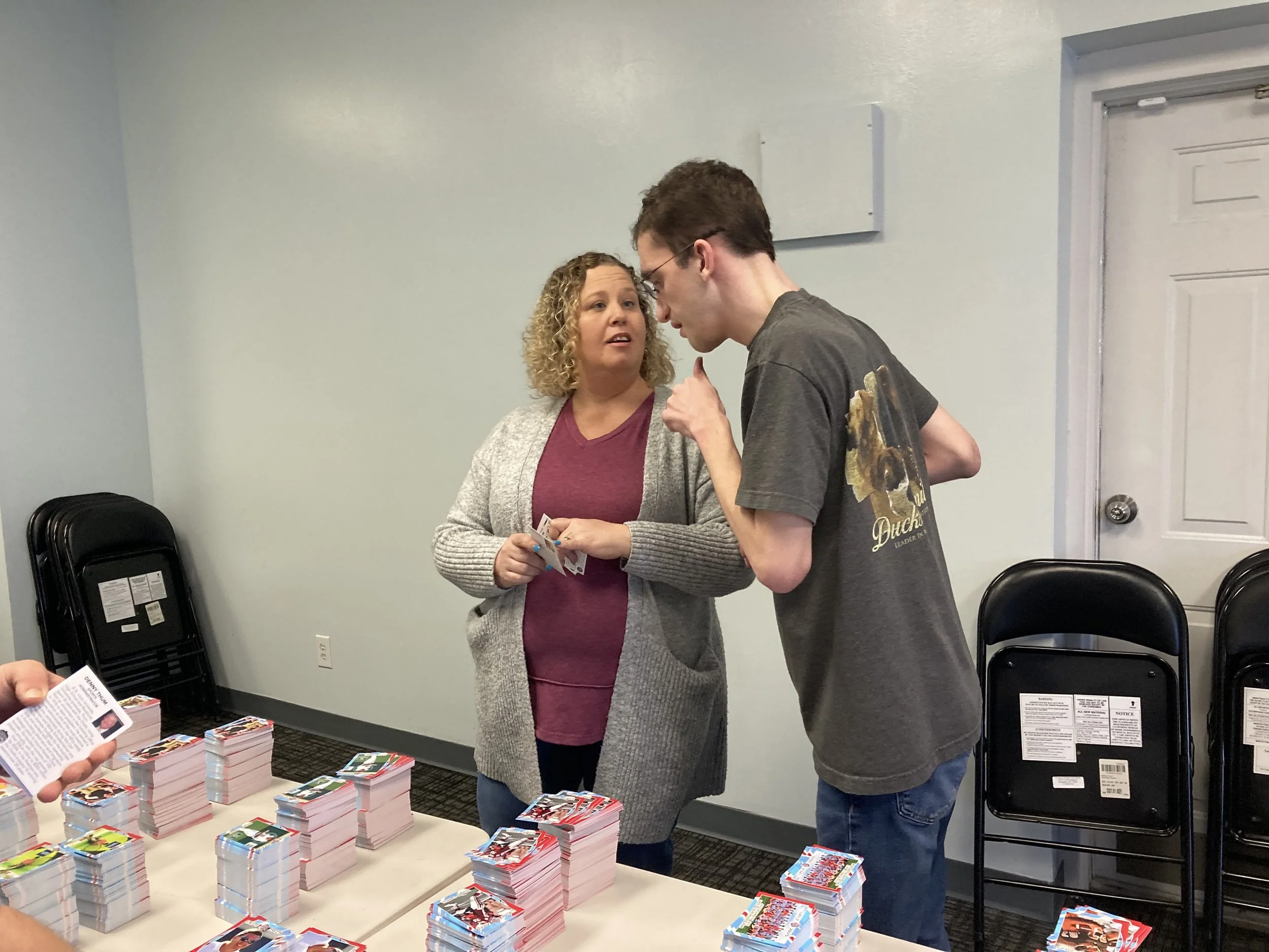 Two people are engaging in an exchange at a table with stacks of trading cards or stickers. The woman has curly blonde hair and is wearing a maroon shirt and gray cardigan, while the young man has short brown hair, glasses, and is wearing a gray T-shirt with a graphic. They are indoors in a room with chairs and light-colored walls.