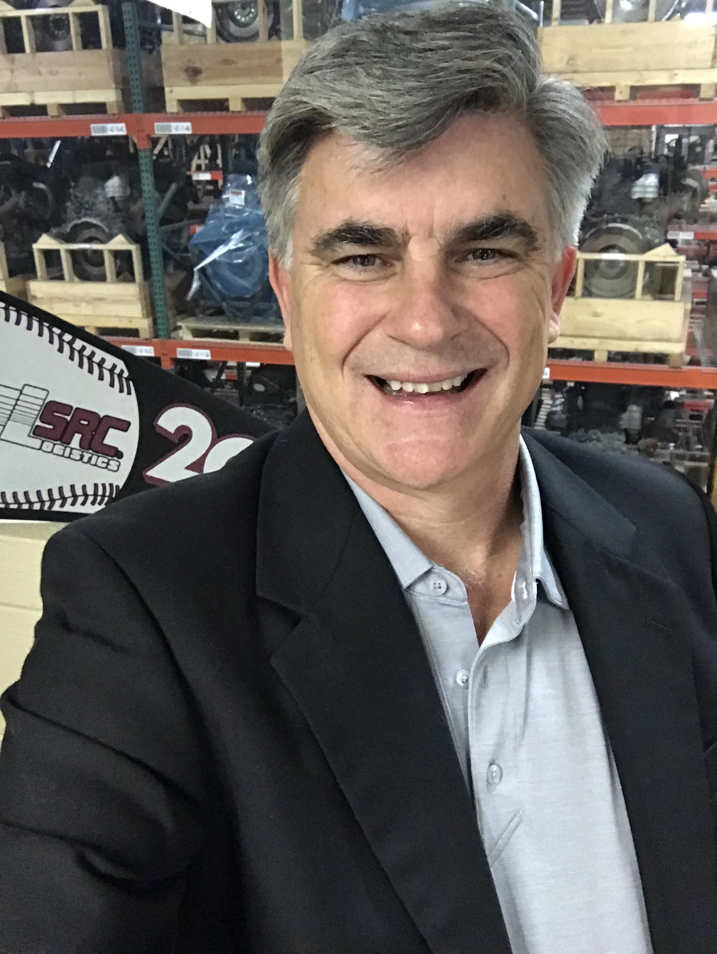 A man in a black blazer and light gray button-down shirt smiling at a camera. In the background, there are shelves with boxed mechanical parts or machinery in a warehouse setting.