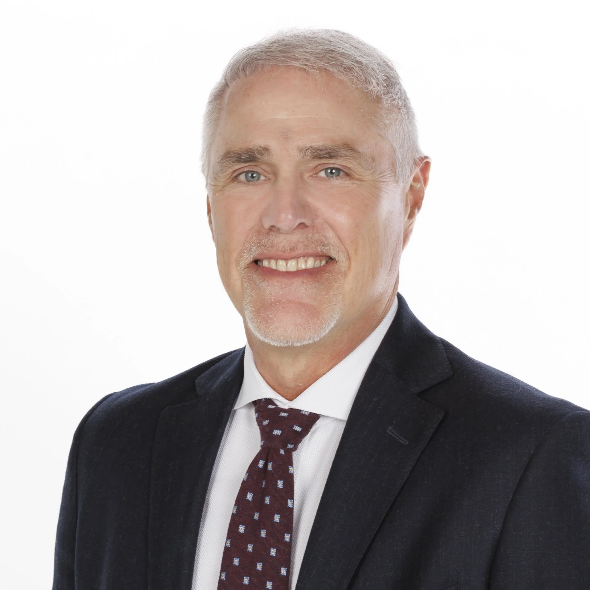 Headshot of a middle-aged man with gray hair, blue eyes, and a light beard, wearing a dark suit, white shirt, and a patterned tie against a plain white background.
