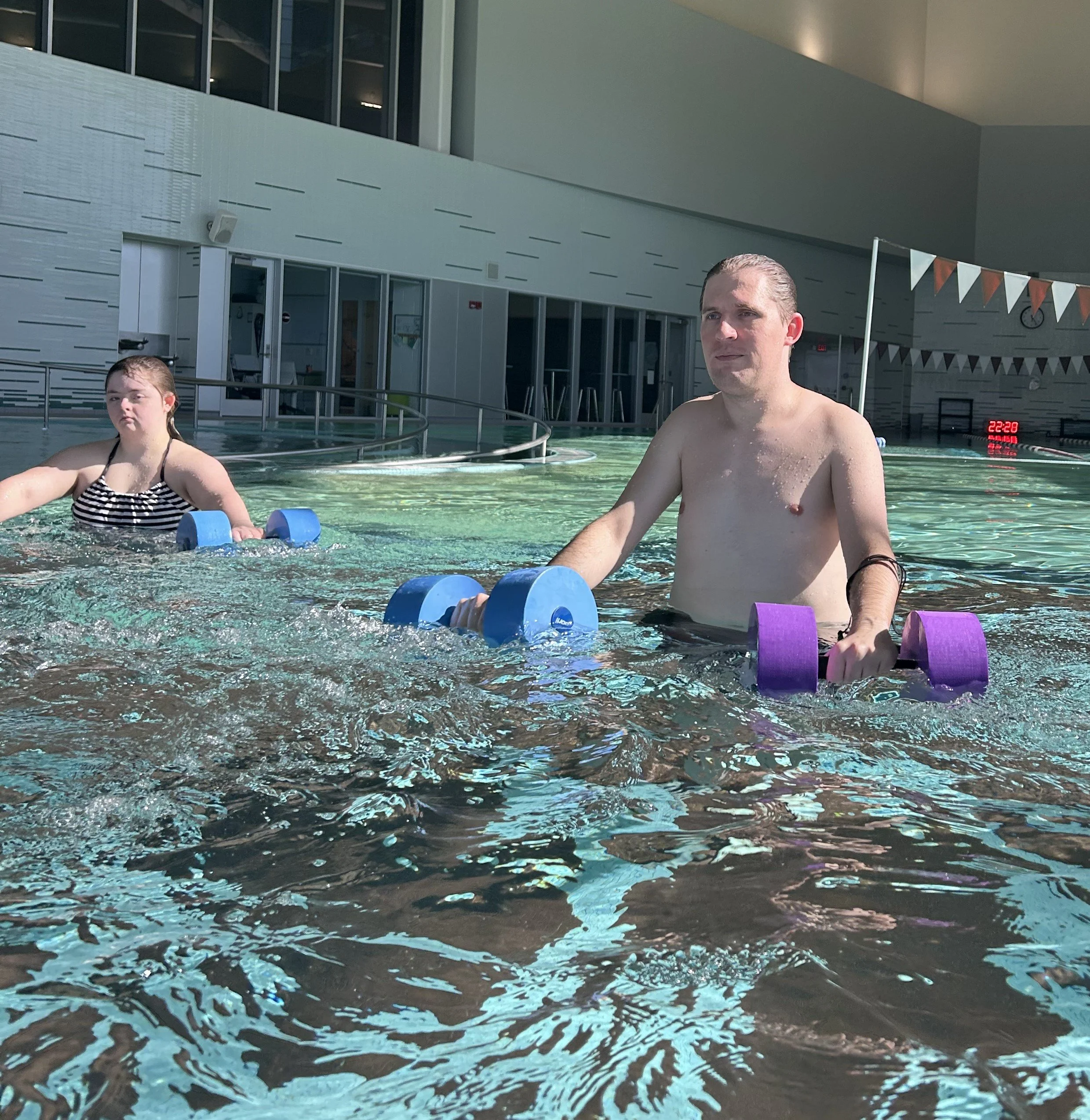 Indoor swimming pool with two people in the water, a woman and a man, and pool safety equipment, with a modern building interior and a digital clock displaying 10:28 PM.
