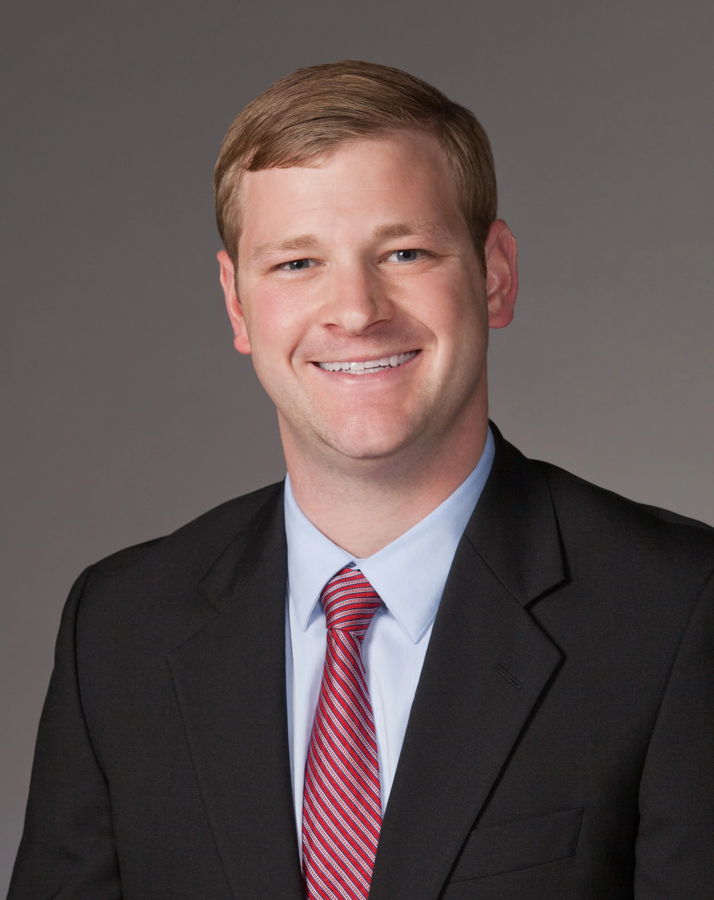 A professionally dressed man in a black suit, white shirt, and red striped tie, smiling against a plain gray background.