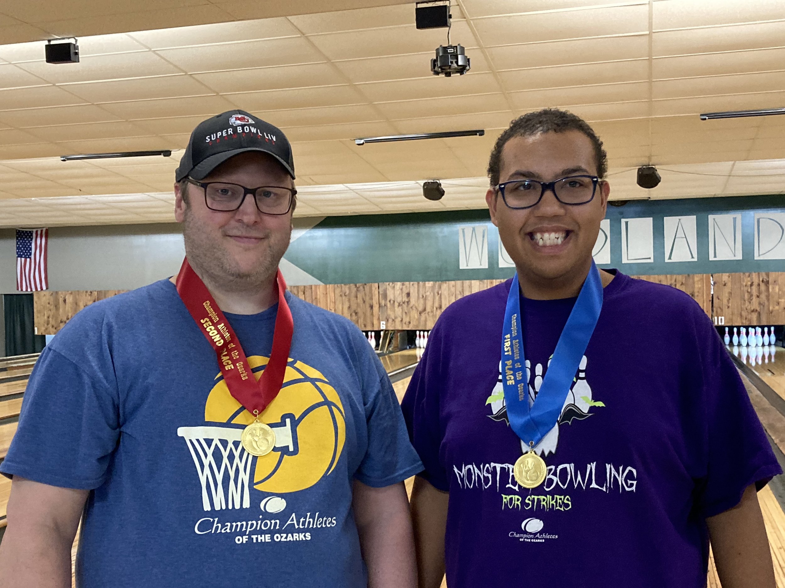 Two men wearing glasses, medals, and champion T-shirts standing in a bowling alley with a wooden floor and pins in the background.
