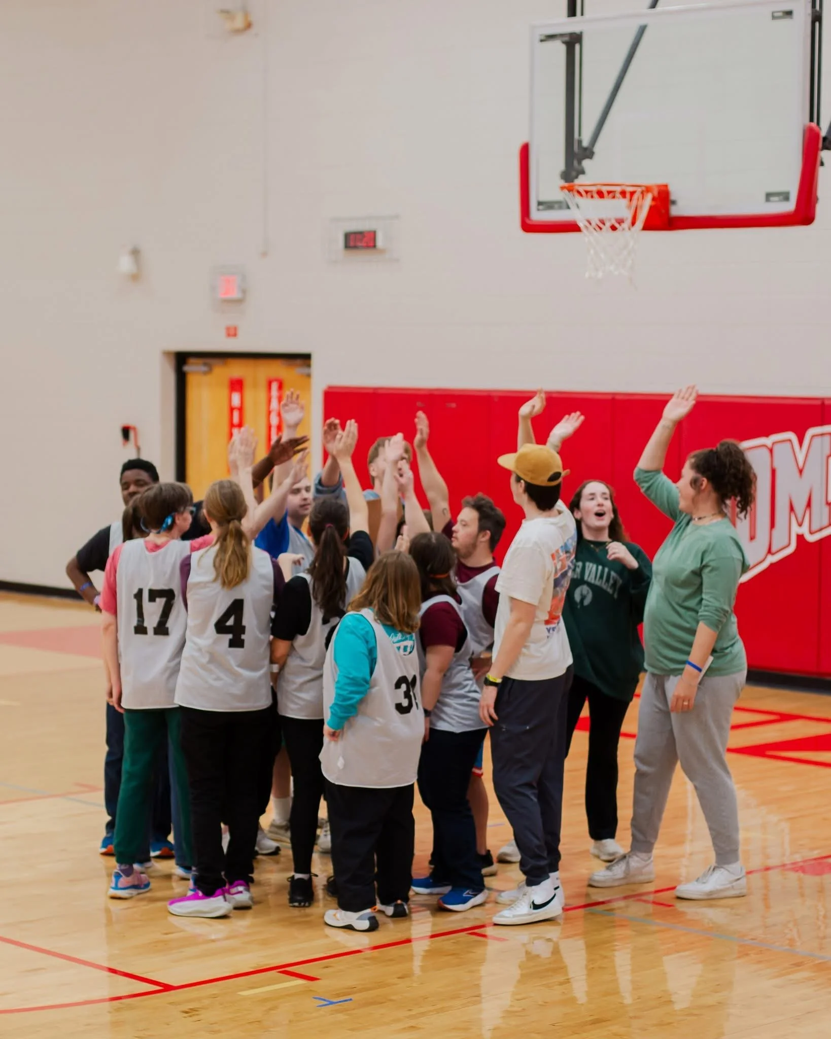Group of people gathered in a gymnasium, some wearing numbered sports jerseys, raising their hands, possibly during a team activity or celebration. A basketball hoop and backboard are visible on the wall behind them.
