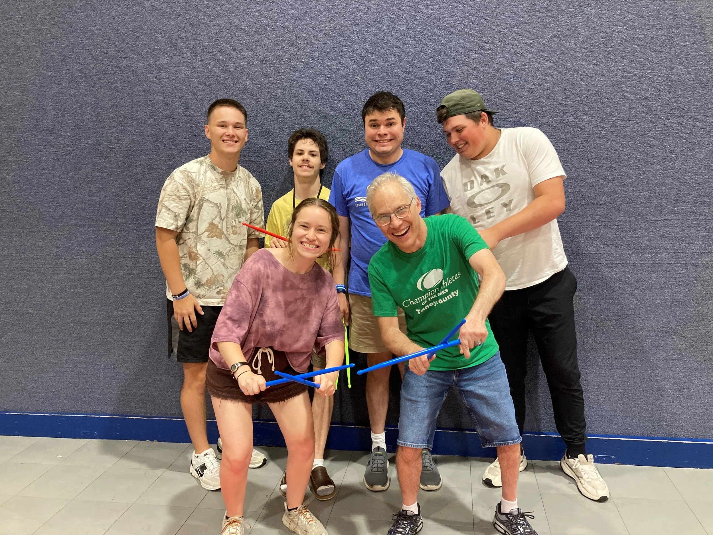 Group of six people, smiling and posing together against a blue textured wall, with one person holding a blue stretch band.