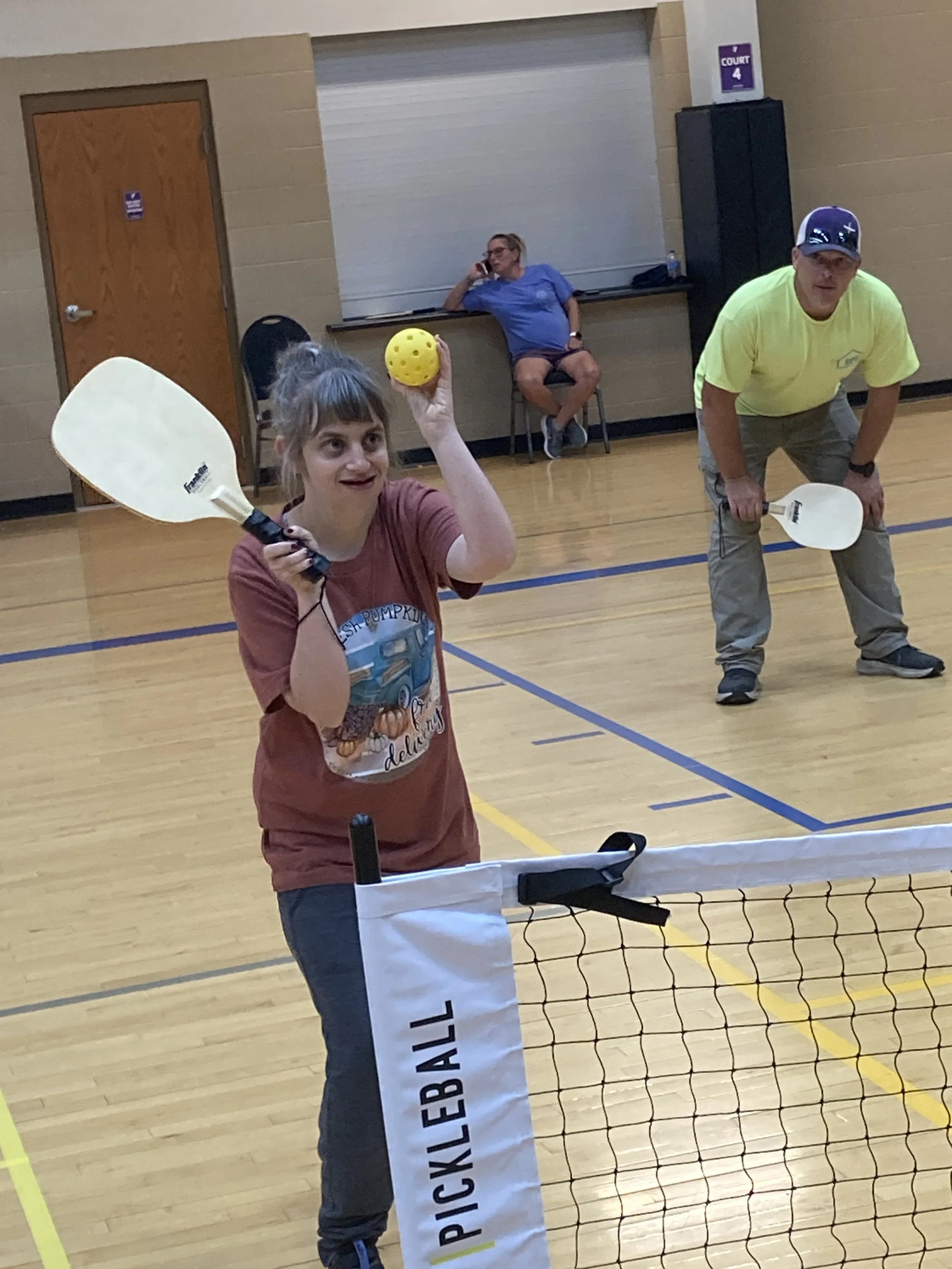 A girl holding a yellow ball and a paddle, preparing to hit the ball during a pickleball game at an indoor sports facility, with a net and a court sign in the background.