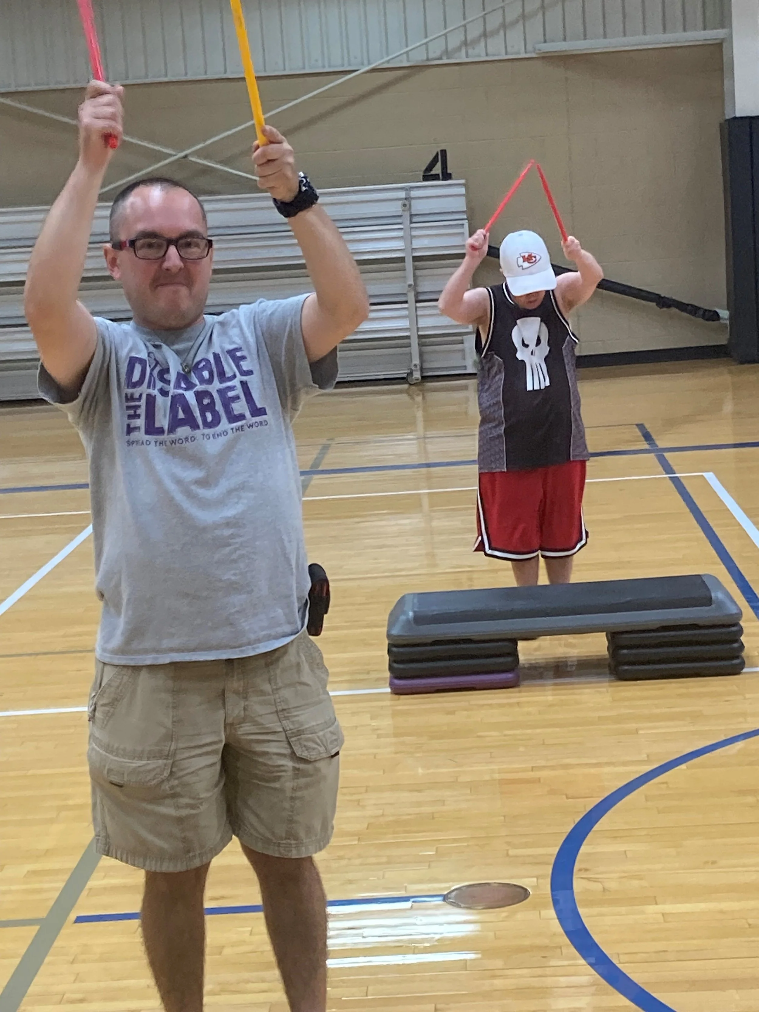A man wearing glasses, a gray t-shirt, and beige shorts standing in a gymnasium with wooden floor, holding foam sticks. In the background, a person wearing a baseball cap, black shirt with a skull design, and red shorts, is also holding foam sticks, with gym bleachers and a wall with a number four visible.