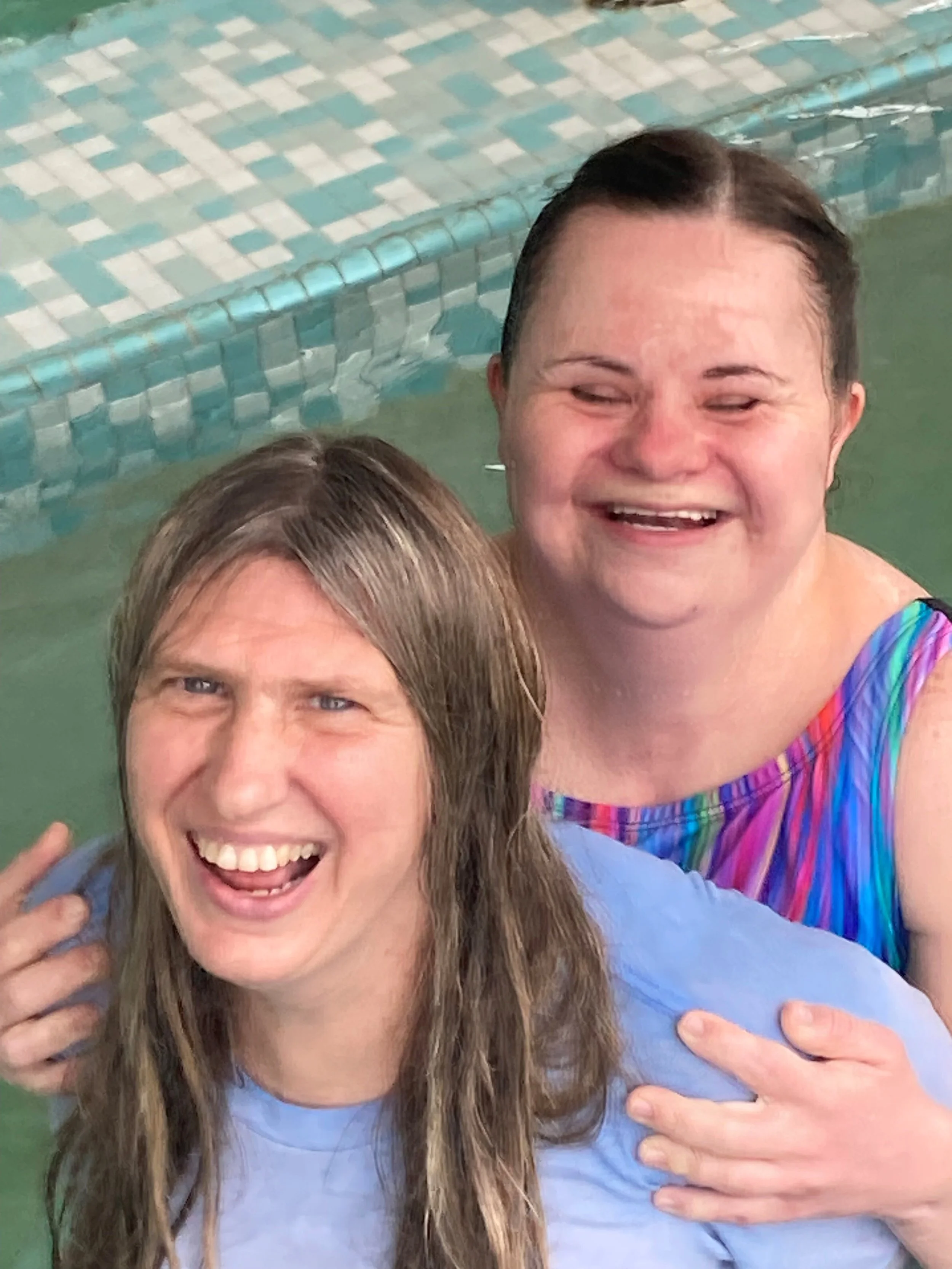 Two women smiling and enjoying time together in a swimming pool, with one woman wearing rainbow-colored clothing.