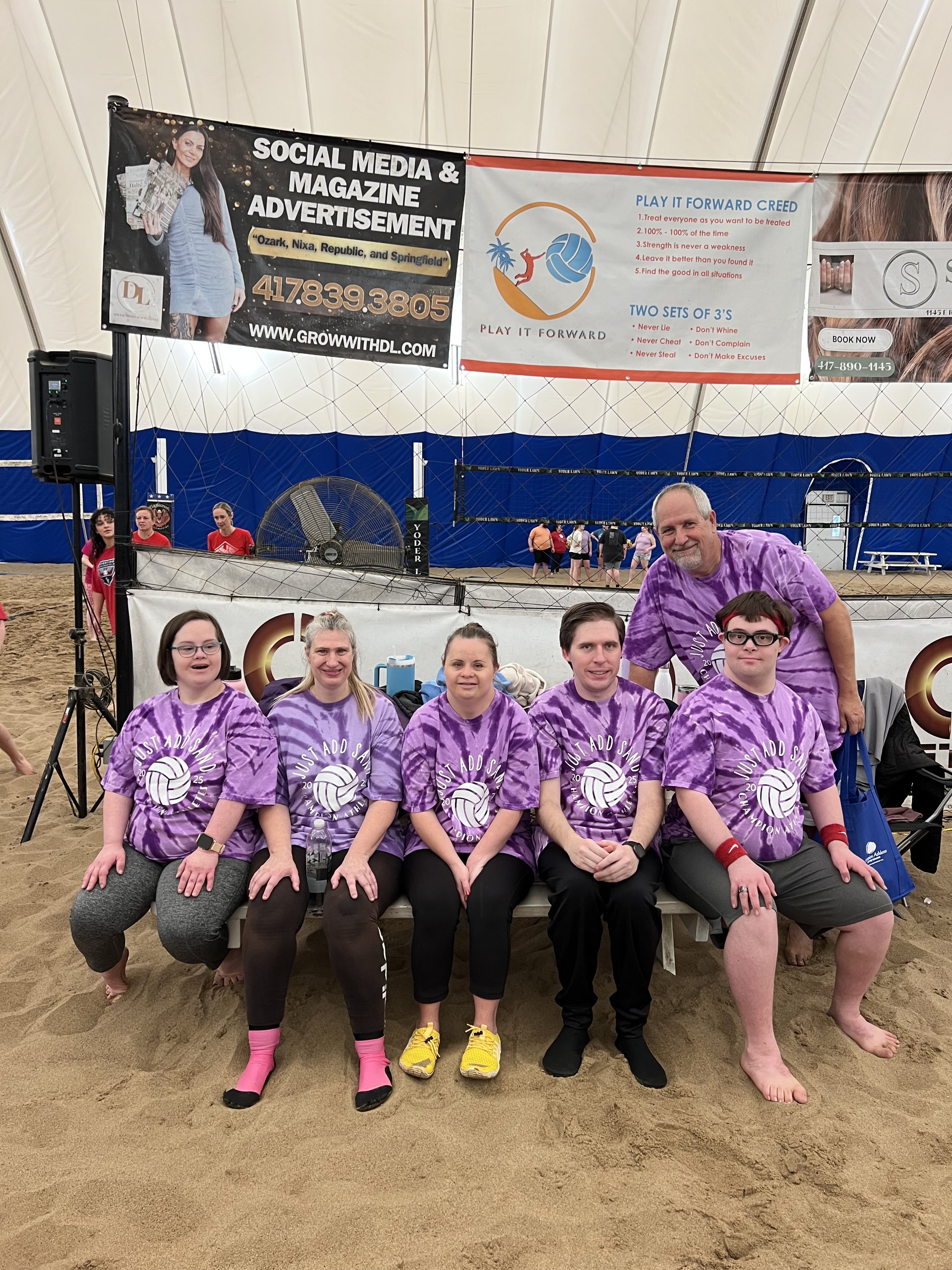 Group of people sitting and standing on sandy floor inside a sports facility, some wearing purple tie-dye T-shirts with a volleyball logo, others standing and smiling, with banners and volleyball net in the background.