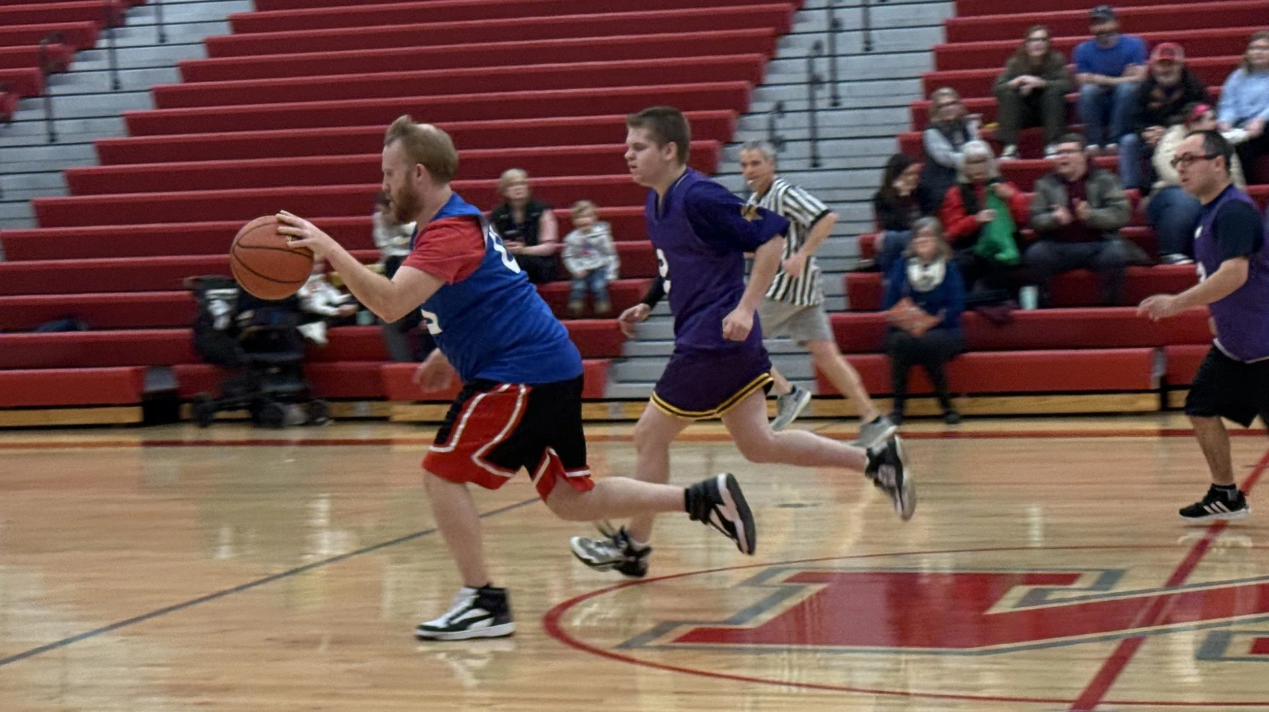 People playing basketball in an indoor gym, with spectators seated in red bleachers in the background.