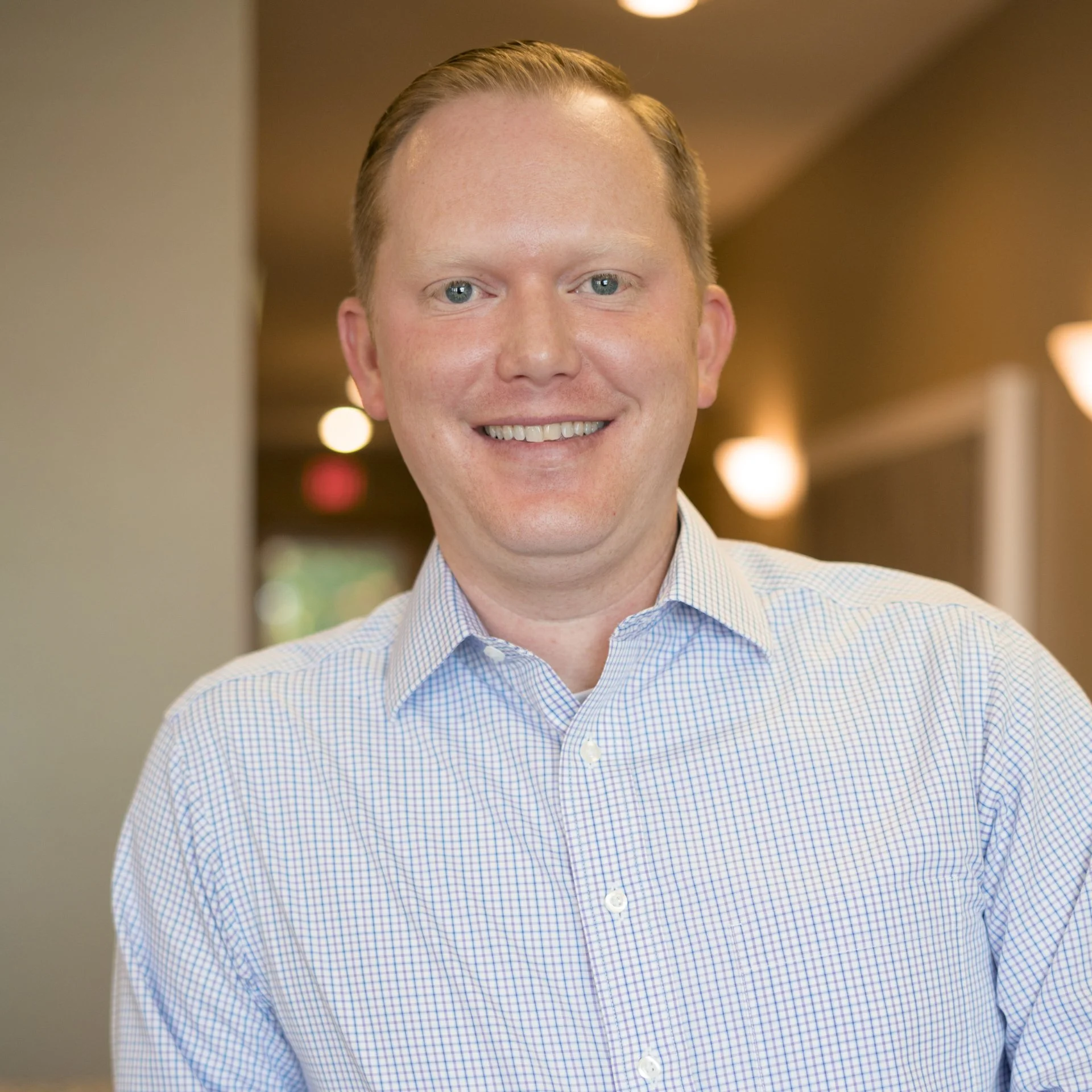 Portrait of a smiling man with red hair, wearing a light-colored checkered shirt, indoors with blurred warm lighting in the background.