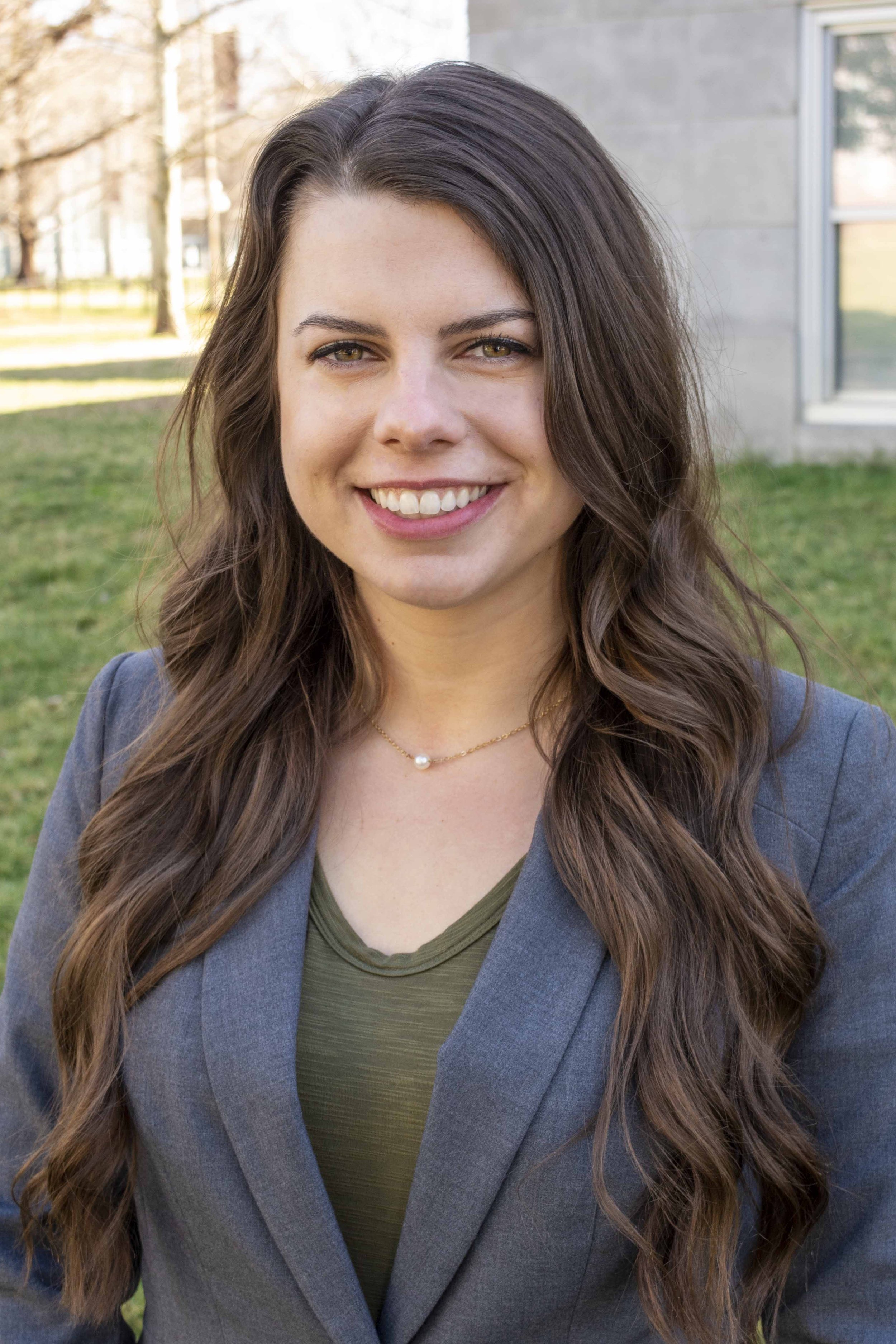 Close-up of a smiling woman with long wavy brown hair, wearing a gray blazer and a green top, outdoors with grass and trees in the background.