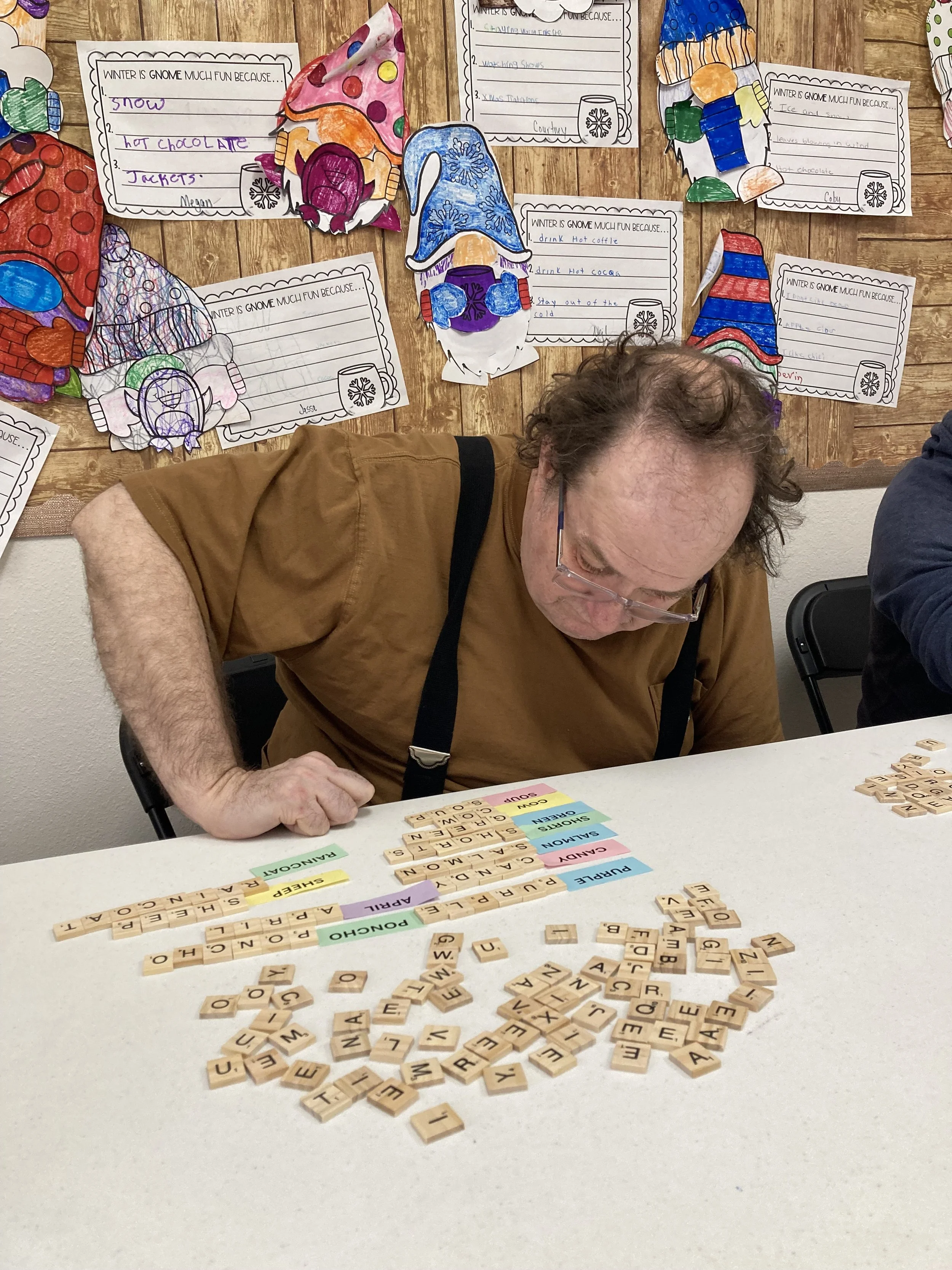 A man is playing a word game with Scrabble tiles on a white table. The tiles spell out words like 'PURPLE,' 'SHORE,' and 'PUNCH.' There are also colored tabs with different categories on the table. In the background, there is a bulletin board with colorful paper illustrations of wrapped candies and handwritten notes.