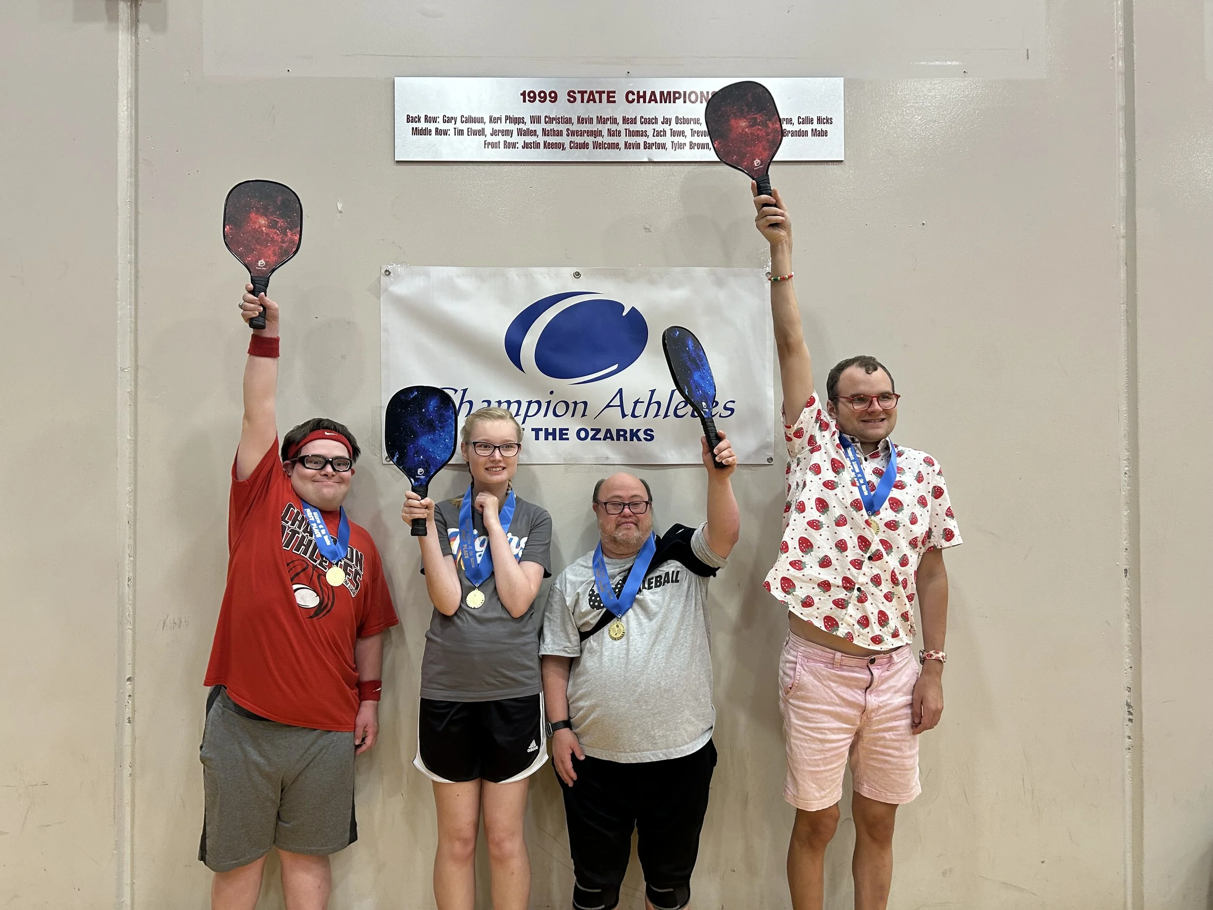 A group of five people celebrating and holding paddles at a sports event with a banner reading 'Champion Athletics of the Ozarks' behind them, and a sign showing the 1999 State Champions list.