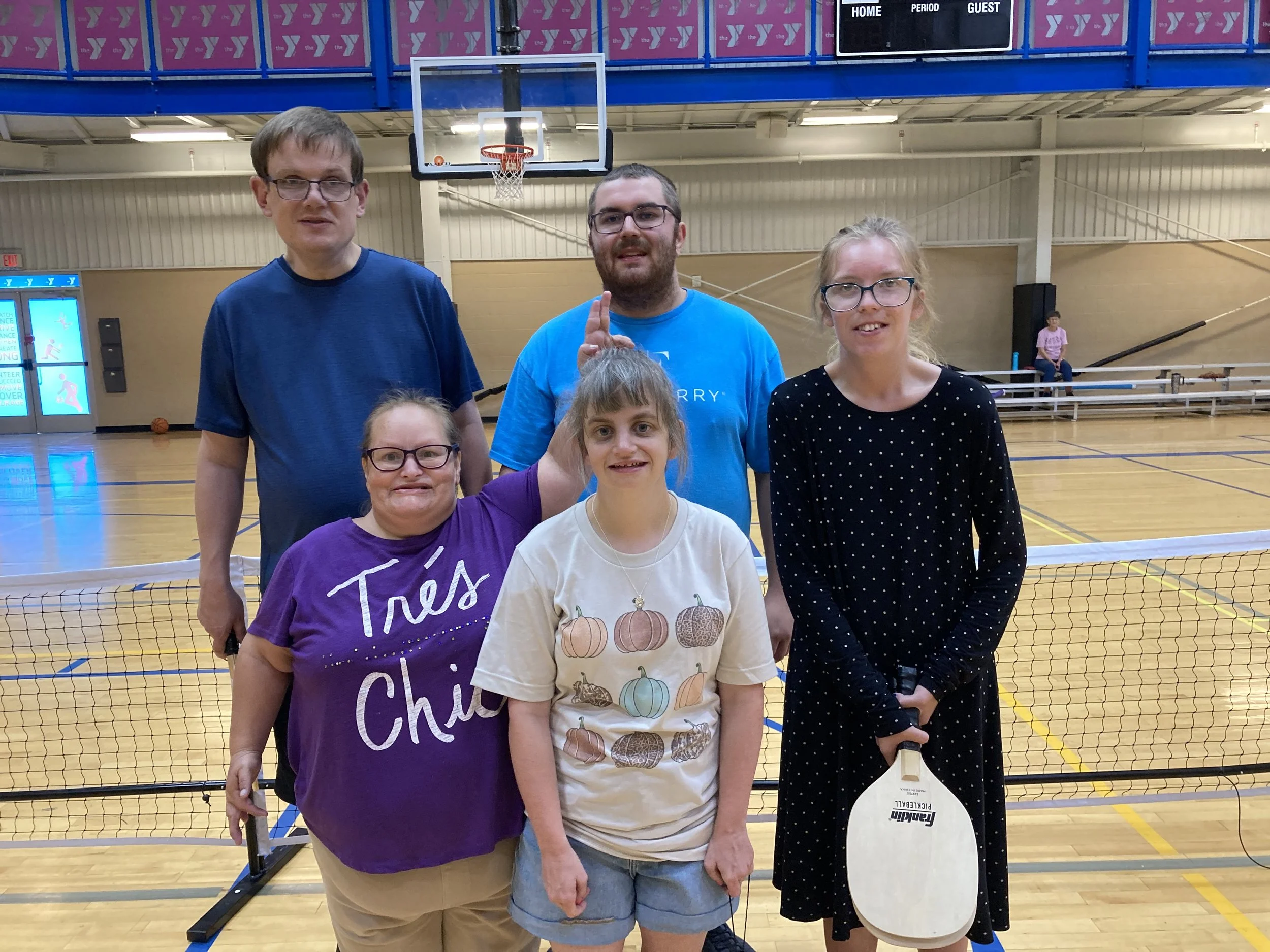 A group of six people, three adults and three children, are standing in a gymnasium near a volleyball net, posing for a photo. There is a basketball hoop in the background and a digital scoreboard overhead.