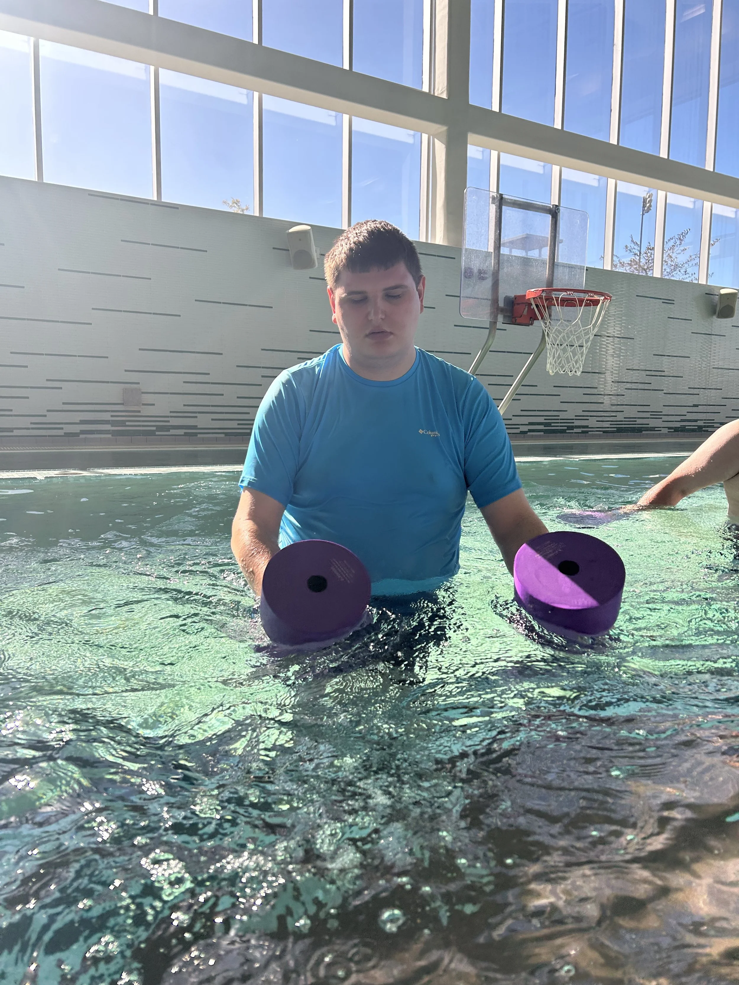 A young man in a blue shirt stands in a pool, holding purple foam dumbbells underwater, with a basketball hoop and large windows in the background.