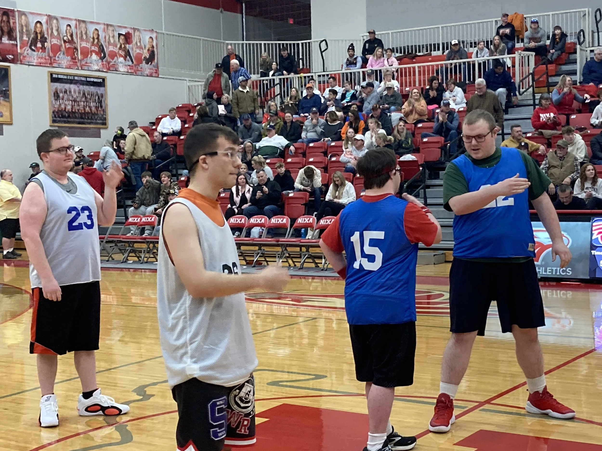 A group of young men in basketball uniforms standing on an indoor basketball court. There are spectators sitting in red bleachers in the background. The gym has basketball posters on the walls and a wooden floor.