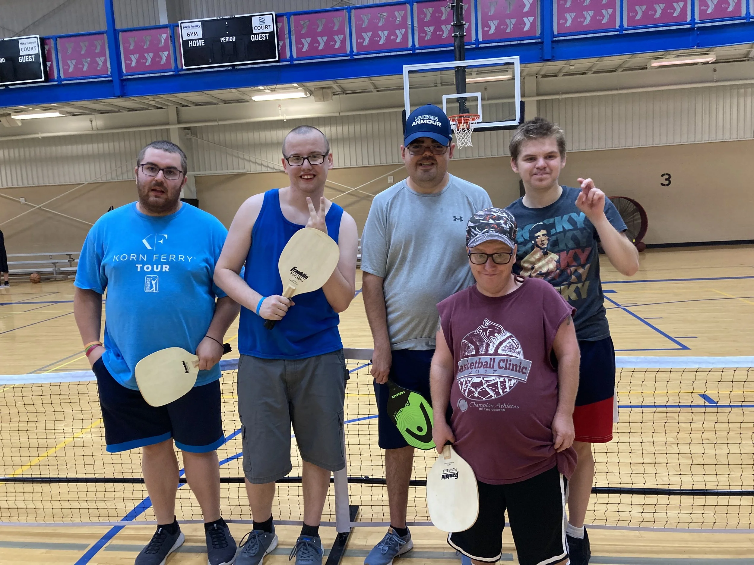 Group of six people standing in a gymnasium with a basketball court, holding pickleball paddles, smiling at the camera.