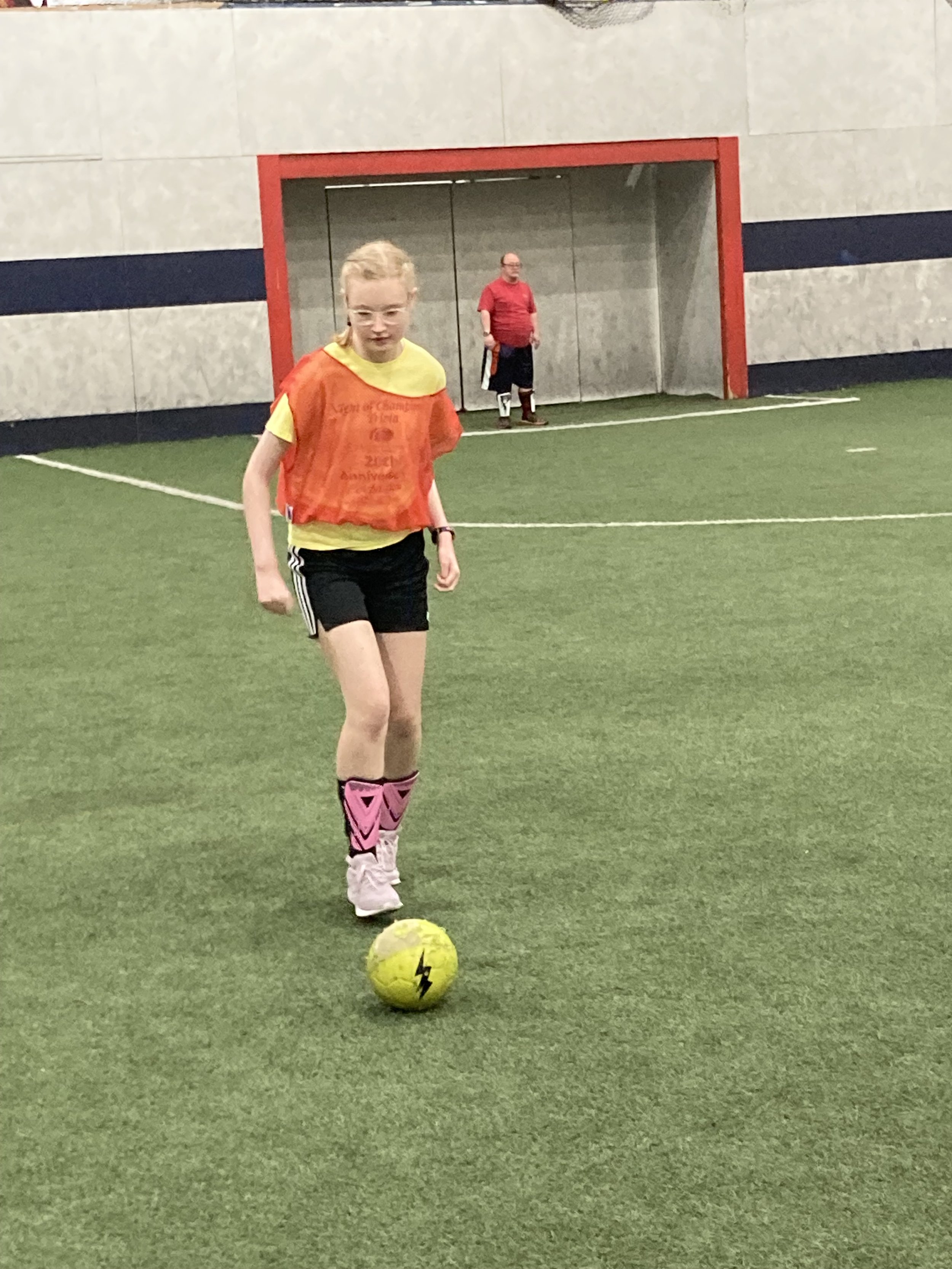 A young girl practicing soccer indoors on a green artificial turf field. She is wearing a yellow shirt, black shorts, and pink soccer socks with cleats. She is about to kick a yellow soccer ball with a lightning bolt logo, while a person in a red shirt and black shorts stands behind her near a goal.
