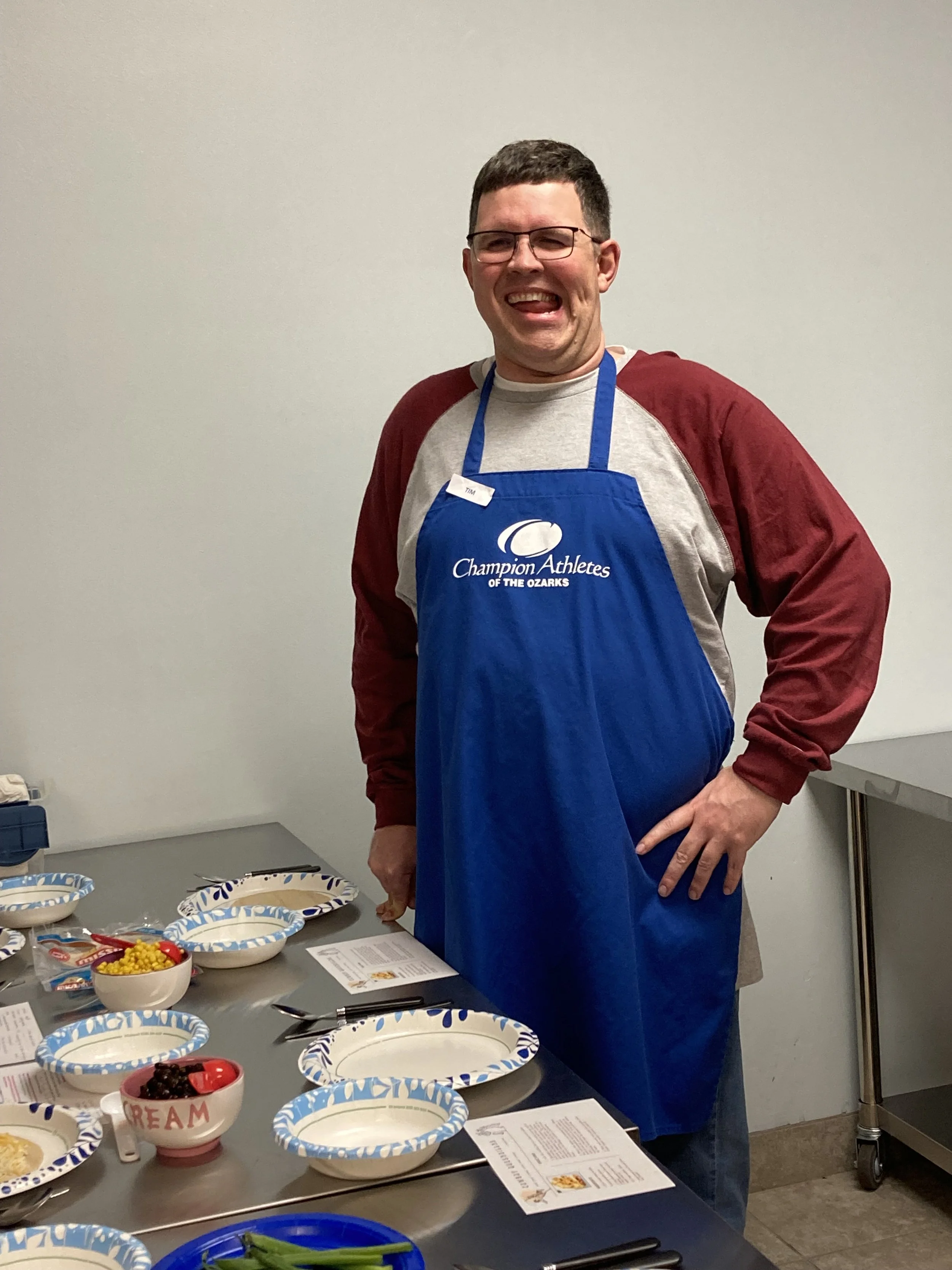 A man with glasses smiling in a room with a table set with bowls of salad, fruit, and papers, wearing a blue apron with the text 'Champion Athletes of the Ozarks' and a name tag that says 'Tim'.