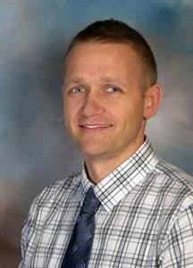 A man with short hair and a friendly smile is wearing a checkered shirt and a tie, standing against a blue and gray background.