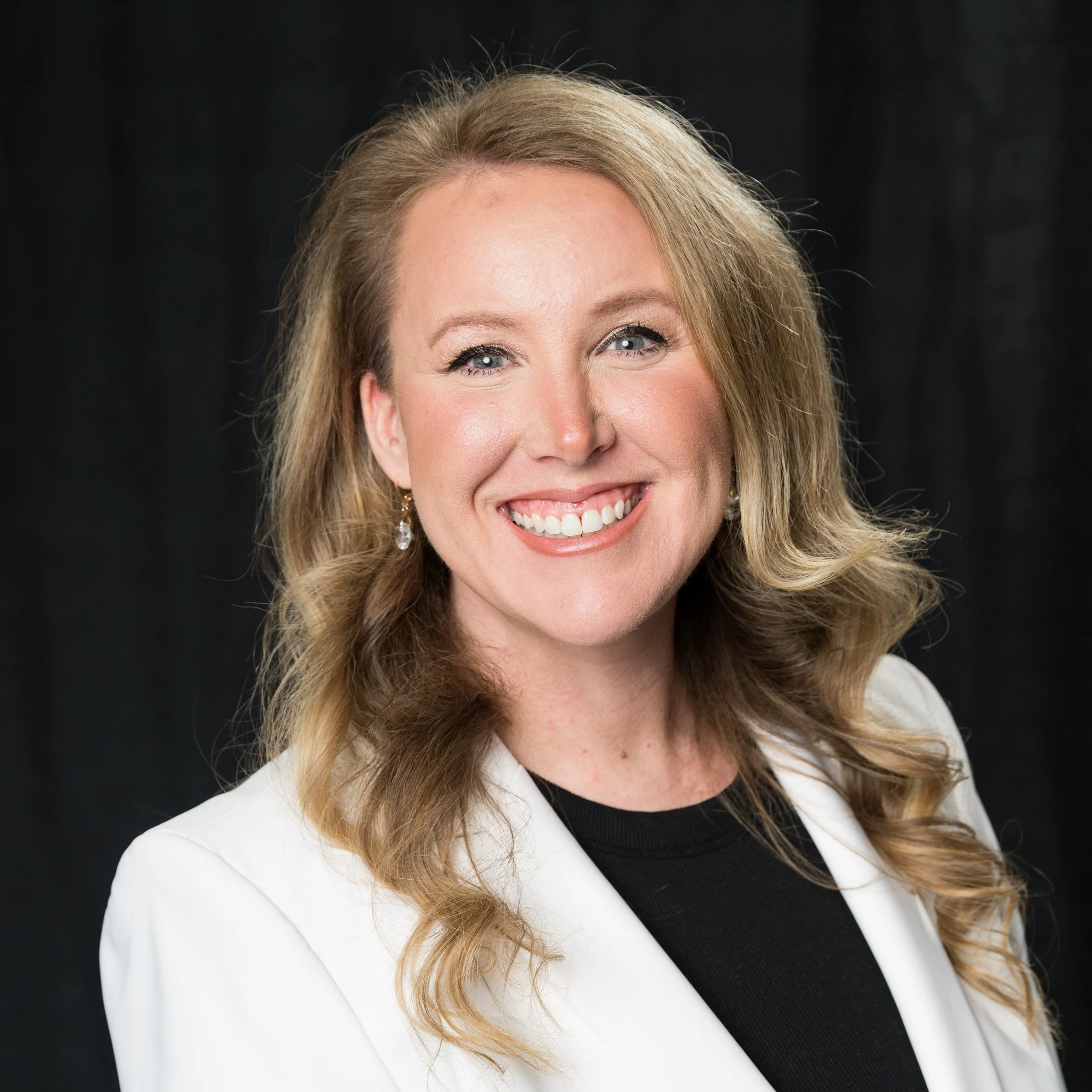 Close-up of smiling woman with blonde hair, wearing a white blazer and black top against a dark background.