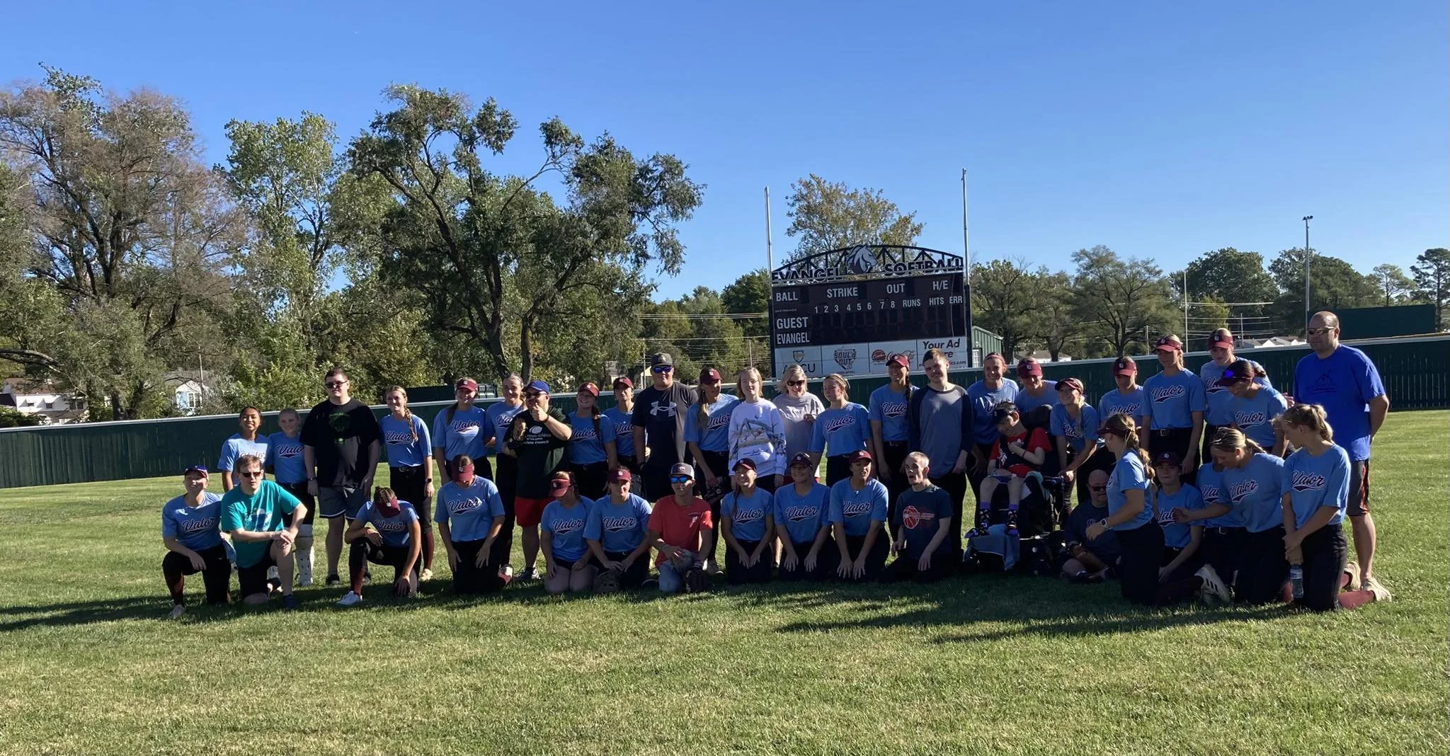 Group of people on a grassy field, wearing athletic clothing, with trees and a sports scoreboard in the background on a sunny day.
