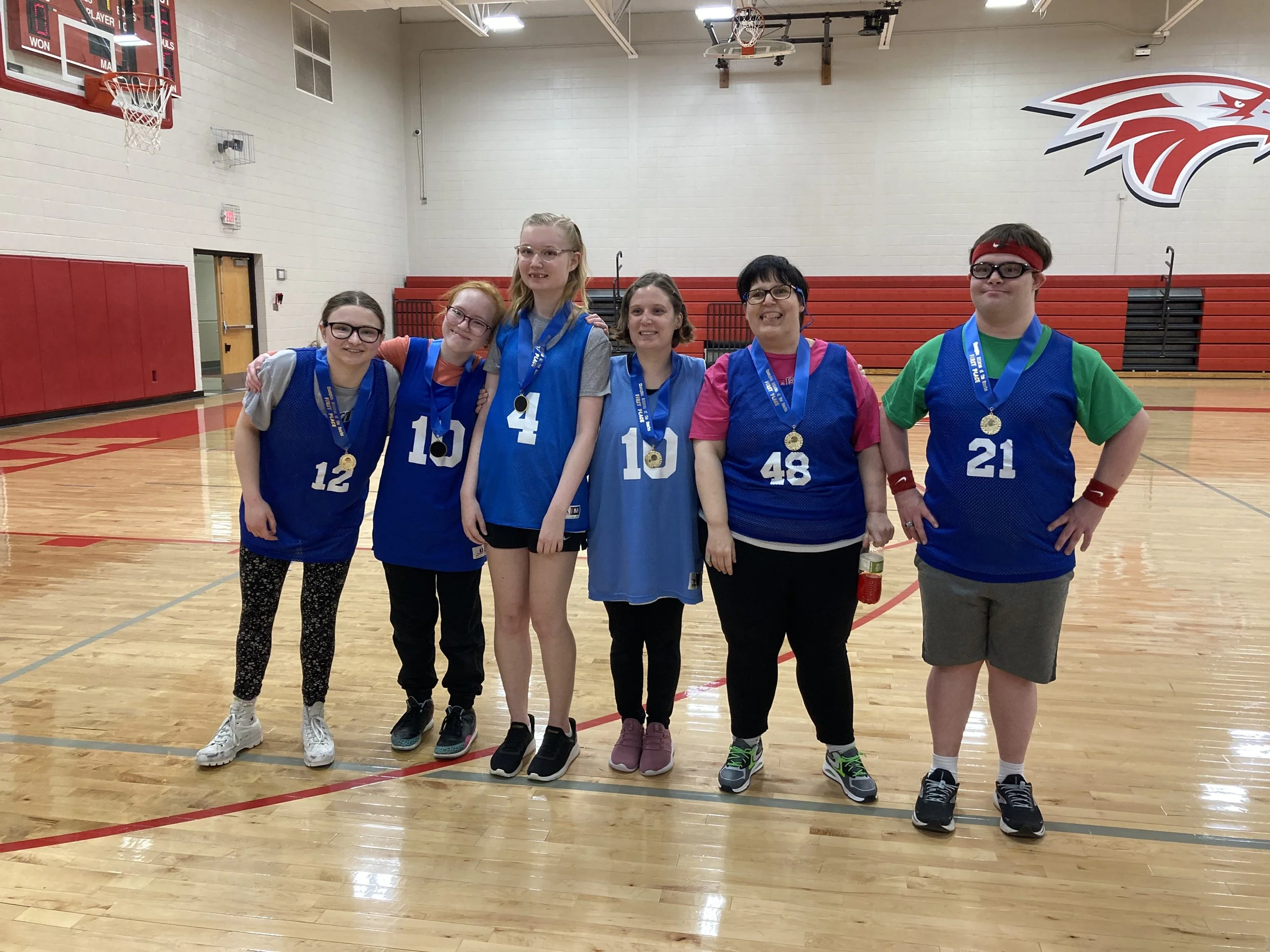 Group of six individuals standing in a basketball gymnasium, wearing medals and sports jerseys, celebrating after a basketball game.