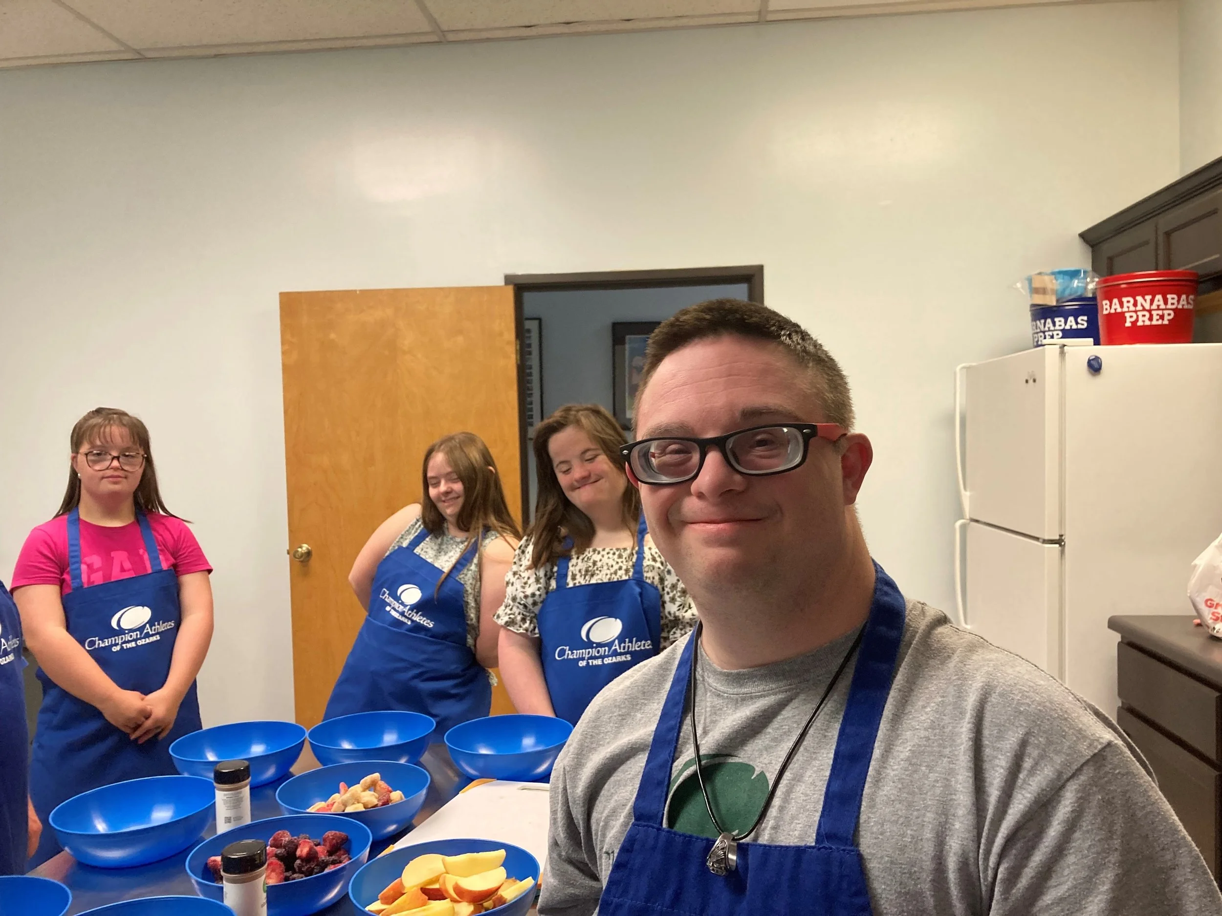 A group of people, primarily young girls, wearing blue aprons with a logo that says 'Champion Athletes of the Ozarks,' gathered in a kitchen or classroom setting. One man in the foreground is taking a selfie, smiling and wearing glasses. On the counter are various bowls filled with fruits such as berries, sliced peaches, and bananas, and containers of spice. There is a white refrigerator with a red and blue container labeled 'Barnabas Prep' in the background.