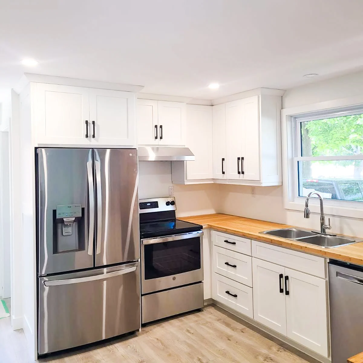 Modern kitchen with stainless steel refrigerator, oven, and dishwasher, white cabinets with black handles, wooden countertop, and a window above the sink showing greenery outside.