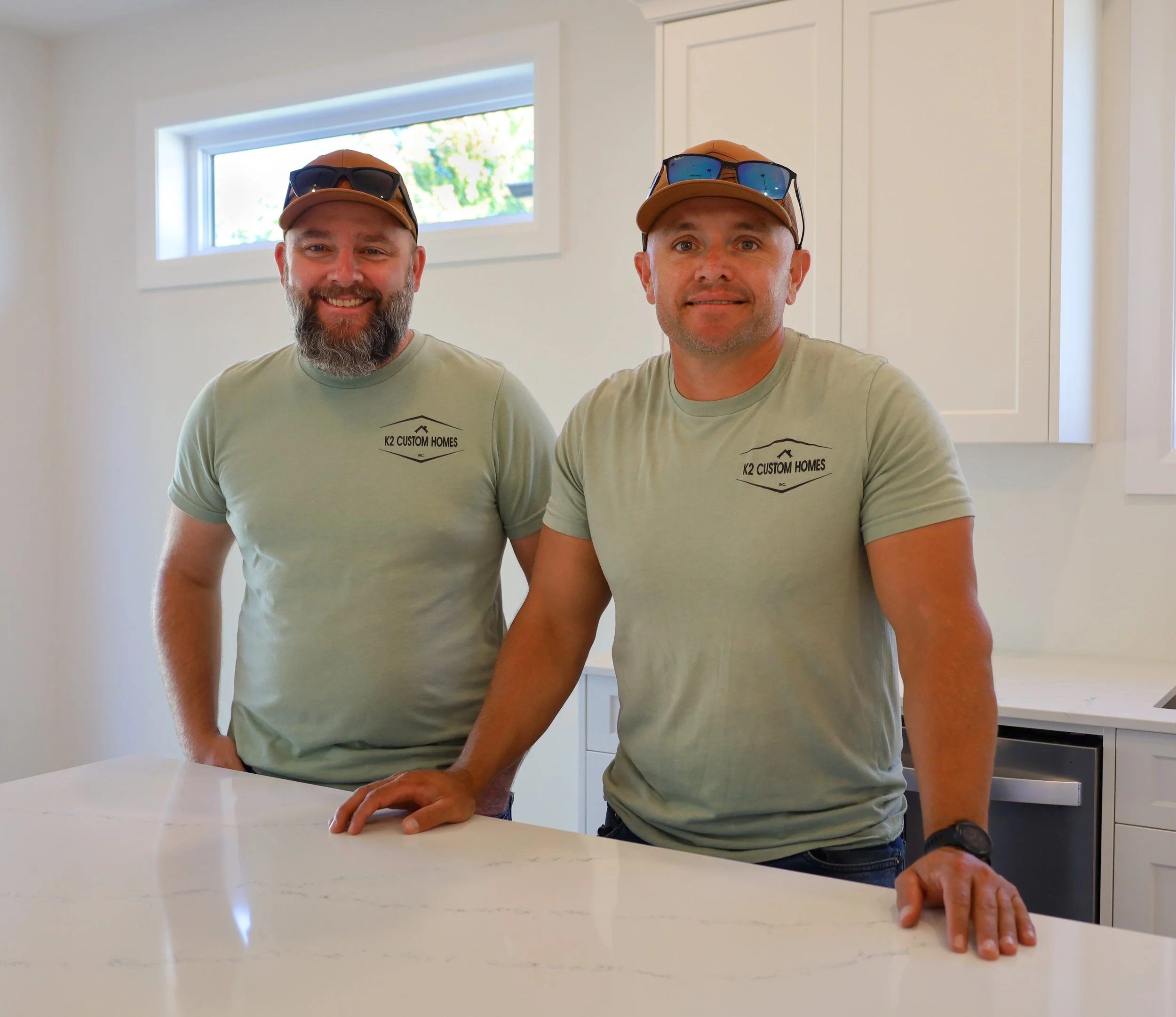 Two smiling men in matching light green t-shirts and caps with sunglasses, standing behind a white kitchen counter in a modern kitchen with white cabinets and a small window.