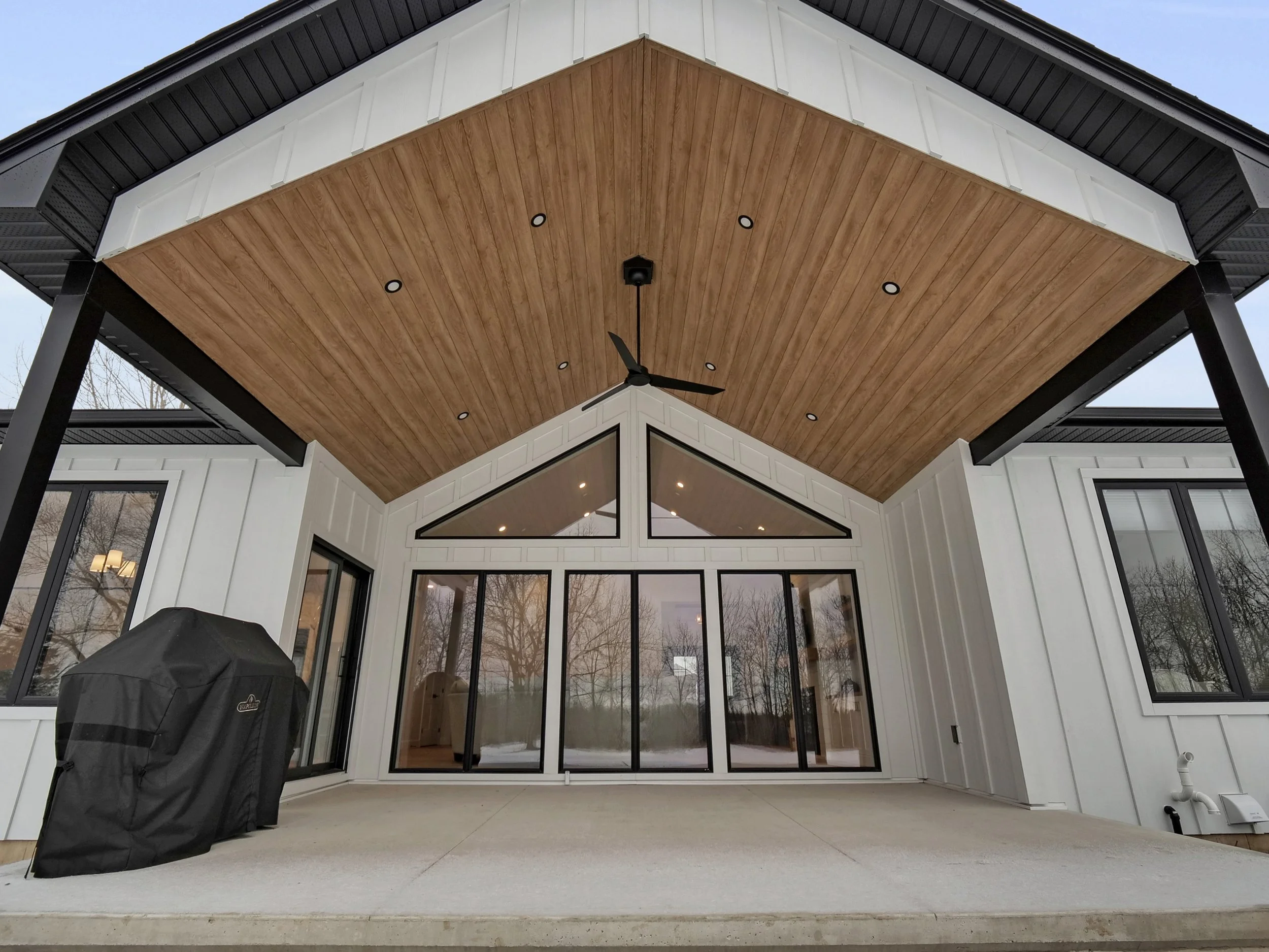 Back porch of a modern house with a wooden ceiling, ceiling fan, large glass sliding doors, and windows, with a covered grill on the left.