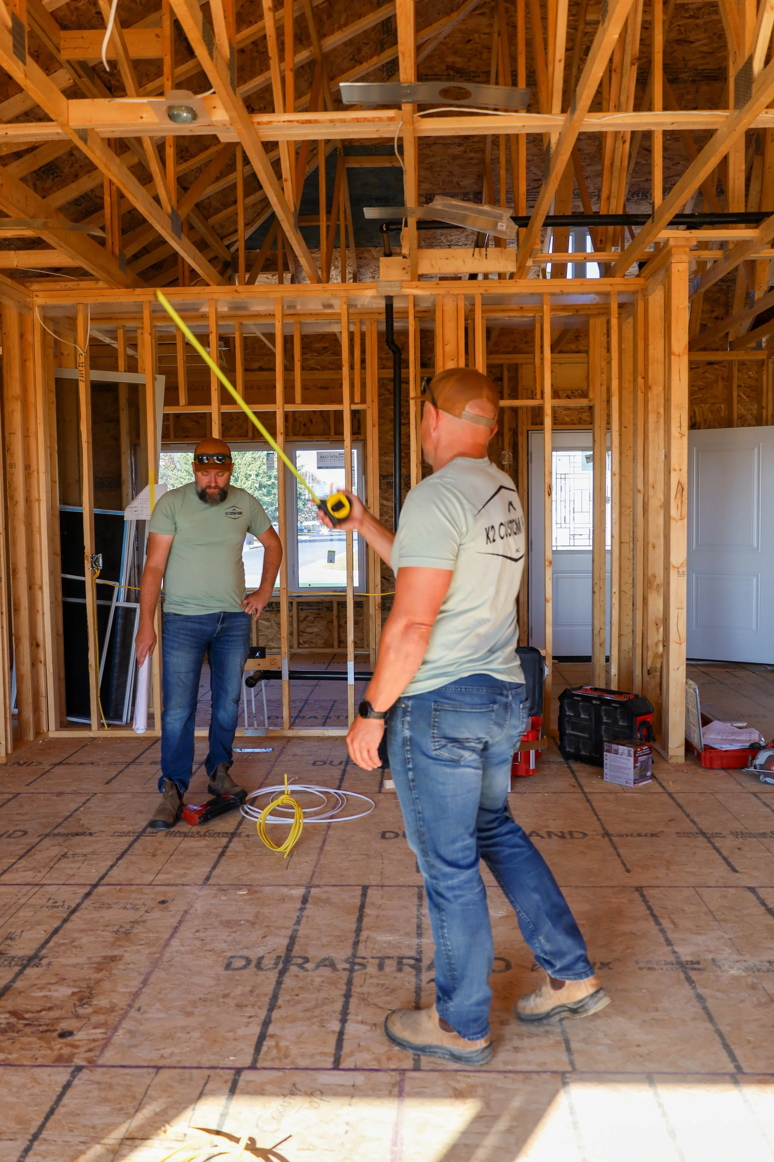 Construction workers inside a house framing, working with electrical wiring and tools, with exposed wooden studs and ceiling. One worker holds a measuring tape, another is standing near wire and tools on the floor.