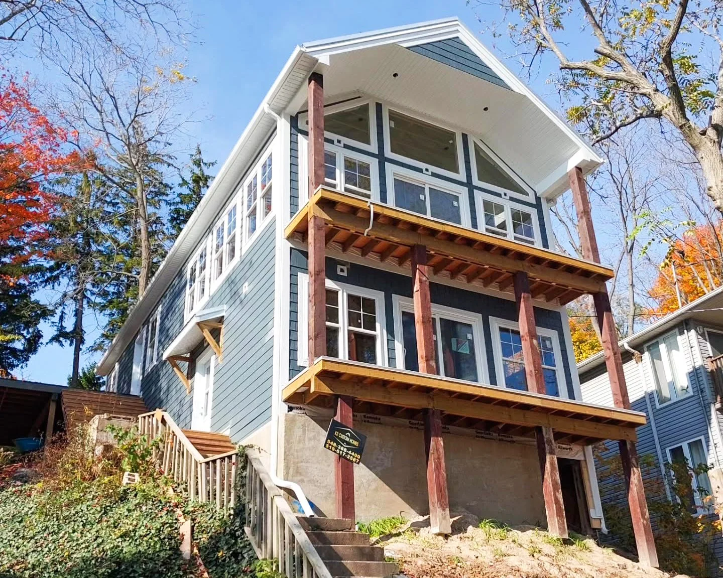 A multi-story house with blue siding and white trim, under construction with exposed wooden support beams on the balconies, situated on a hill surrounded by trees with autumn foliage.